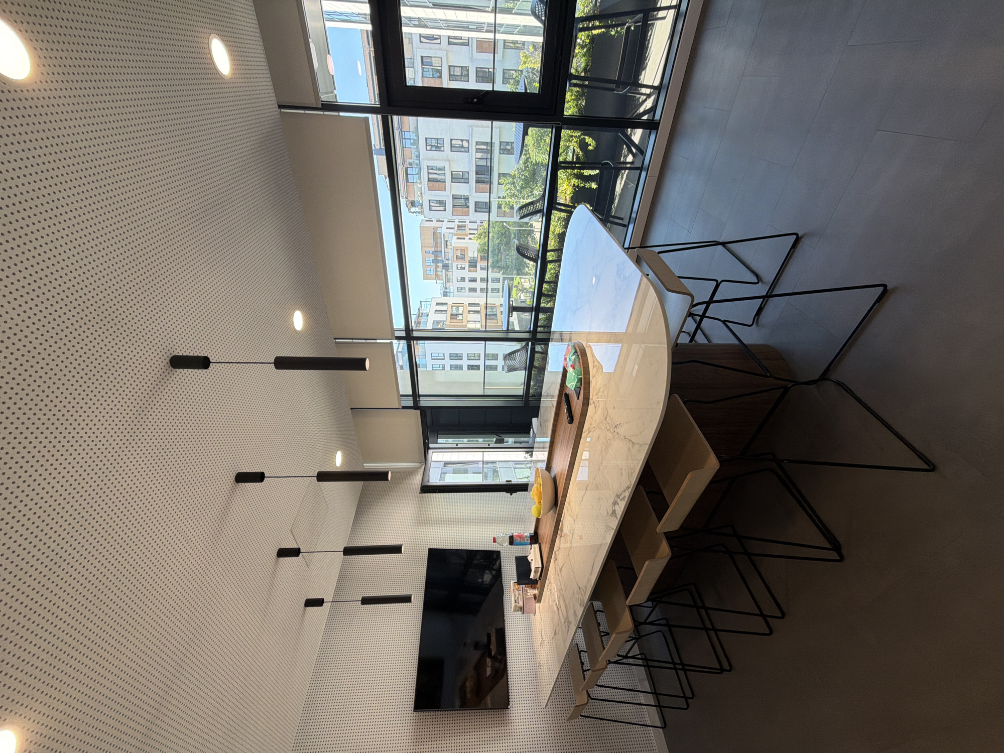 The image shows a modern, communal kitchen and dining area with a long, white marble-topped table. The table has several bar stools with light wood seats and black metal legs arranged around it. On the table, there is a wooden tray with a bowl of lemons, a bottle of water, and some magazines. A large flat-screen television is mounted on the wall behind the table. The room has large windows that look out onto a city skyline with apartment buildings and trees. The ceiling is white with a perforated pattern and features several black pendant lights and recessed lighting fixtures. The floor is a dark gray tiled surface. There are no people visible in the image.
