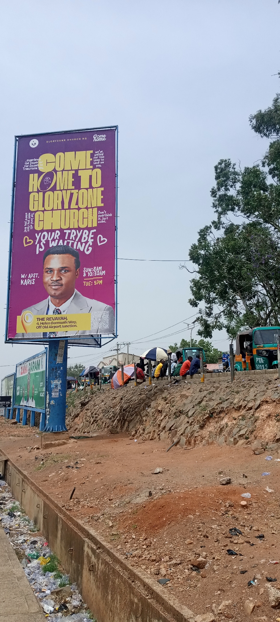 The image shows a street scene in Jos, Nigeria. A large billboard advertising Gloryzone Church HQ dominates the left side of the frame. The billboard is purple and yellow, featuring a photo of a man named W/ APST. Karis. It also lists the church's address: The Revayah, 4, Helen Gomwalk Way, Off Old Airport Junction.
On the right side of the frame, a rocky embankment rises above the street level. Several people are sitting or standing on top of the embankment, some under umbrellas. In the background, there are commercial vehicles, including tricycles with the registration number PTA 10715.
A drainage ditch full of trash runs along the bottom left of the frame. The ground is red dirt. The sky is overcast.