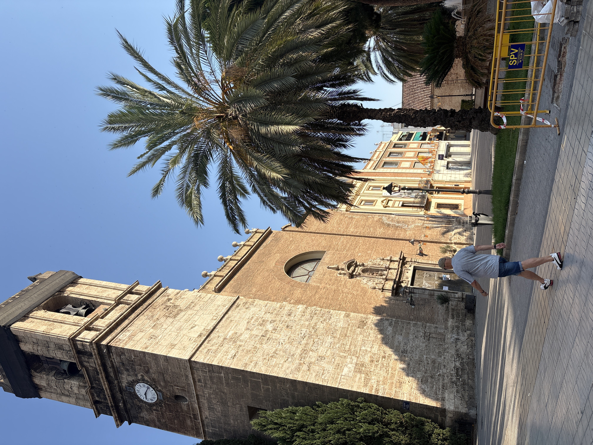 In Valencia, Spain, on a clear day, a man walks on a paved walkway in front of a large stone and brick building with a tall clock tower. The tower has a clock face visible, and is capped with black netting. Bells are visible through arched openings in the upper section of the tower. The building has architectural details including a large arched window and a decorative stone carving above a doorway. Palm trees are present in the foreground and middle ground, with one tall, mature palm tree dominating the right side of the frame. Behind the main building, other buildings with balconies and awnings are visible, along with a street lamp. A yellow construction barrier with red and white tape is positioned in the lower right corner of the image, with some paving stones and an open manhole cover nearby. The sun casts long shadows across the walkway and building facades.