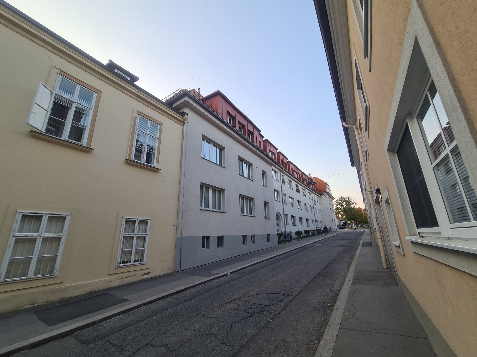 The image depicts a street scene in Vienna, Austria, featuring buildings on either side of a road. On the left, a pale yellow building with white-framed windows, one of which is partially open, occupies the foreground. To its right, a light gray building with numerous windows extends further down the street. This building has a red-tiled mansard roof with dormer windows. A narrow sidewalk runs alongside the buildings, and the asphalt road is cracked. Further down the street, more buildings are visible, and a few pedestrians are walking. The sky is clear and pale blue, suggesting it is daytime. The perspective of the image is from ground level, looking down the street.