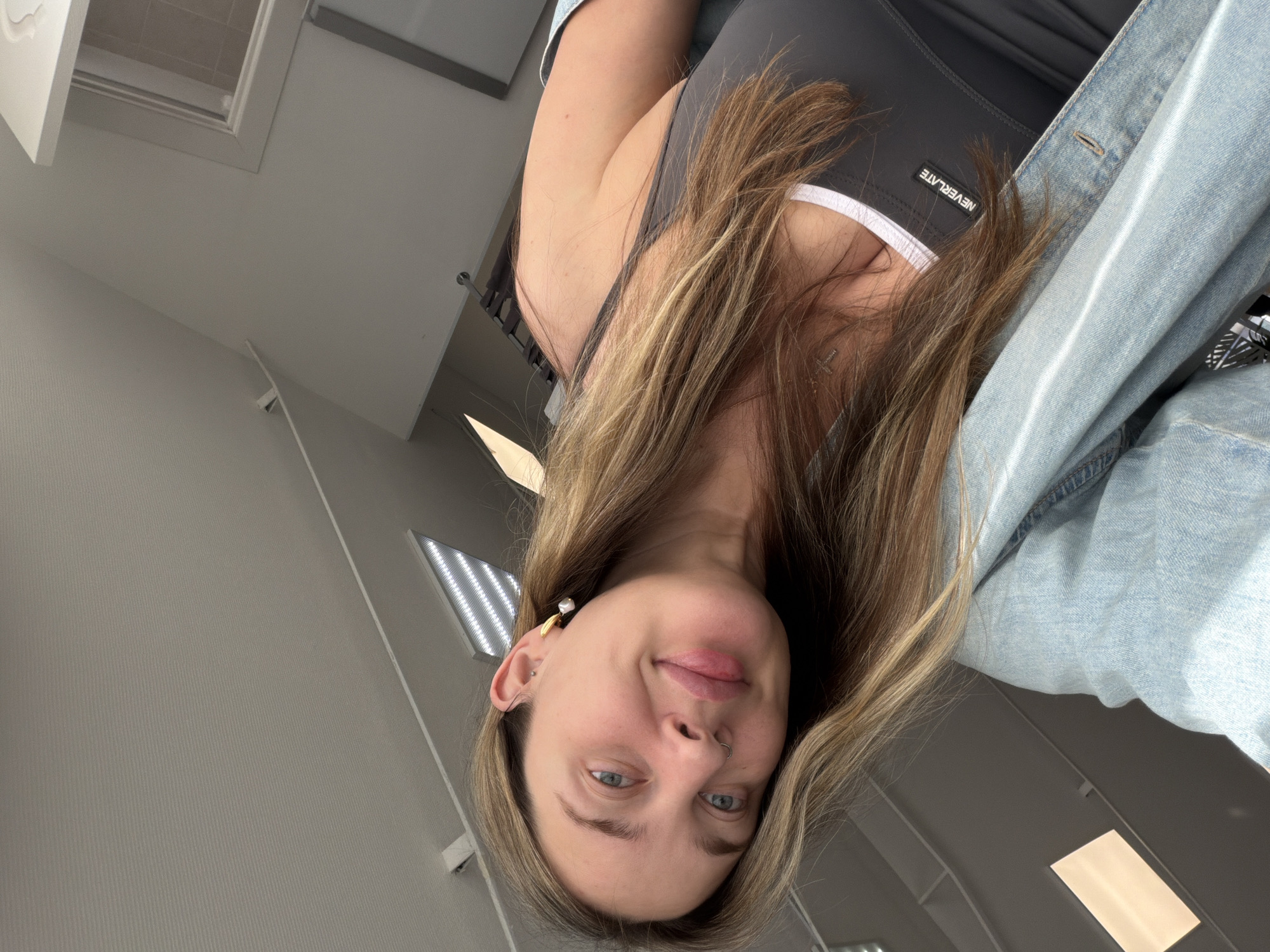 The image captures a young woman from an inverted perspective, likely a selfie, in an indoor setting in Chaikovsky, Russia. Her face, framed by long, light brown hair with blonde highlights, is upside down as she looks directly at the camera with a calm expression and a slight smile. Her blue eyes are open.
She is wearing a dark grey tank top with a white trim along the neckline, over which a light blue denim jacket is draped over her shoulder. The brand name "NEVERLATE" is visible in white lettering on the front of her tank top. Accessories include a small hoop nose ring, a distinctive earring with a gold-colored accent and a white pearl drop on her left ear, and a small cross pendant necklace.
The background consists of light-colored walls and a ceiling with multiple bright linear light fixtures, possibly LED panels or fluorescent tubes. A window or skylight structure is also visible in the upper part of the original image, contributing to the bright indoor lighting.