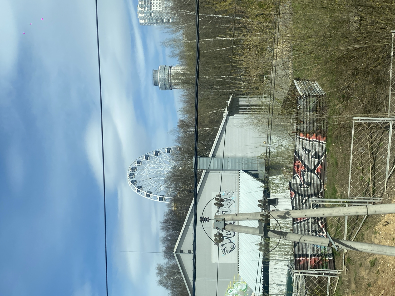 The image displays an urban landscape under a bright, clear blue sky with wispy white clouds. The original image appears to be a portrait photograph, but it is displayed rotated 90 degrees clockwise.
In the mid-ground, a large white Ferris wheel with black gondolas is a prominent feature on the left, partly surrounded by bare trees, suggesting an amusement park setting. To its right, a distinctive multi-story cylindrical building with numerous windows and a rounded top, possibly an observation tower, stands tall. Further in the background, a modern, tall apartment building with many windows rises towards the sky.
The foreground is dominated by thick black power lines running diagonally across the frame and a utility pole with insulators. Below these lines, several structures are visible. A light-colored building with a corrugated metal roof on the lower left has stylized graffiti on its side, including bubble letters that could spell "POG" or similar. Adjacent to it, a longer, low structure made of corrugated metal sheets is heavily covered in graffiti, featuring black, white, and red stylized letters that resemble "SKR." A chain-link fence borders the scene on the lower right. The ground throughout the foreground and mid-ground is covered in sparse, brown vegetation, indicating early spring or late autumn. High in the upper left corner of the sky, three small, pink, heart-shaped balloons are floating.
The scene depicts a daytime setting with bright sunlight. No people are visible, so no specific activity or emotions are discernible, but the presence of the Ferris wheel points to a recreational area. The blend of modern buildings, amusement park features, and graffiti suggests an urban environment, likely in Khimki, Russia, as per the location context.