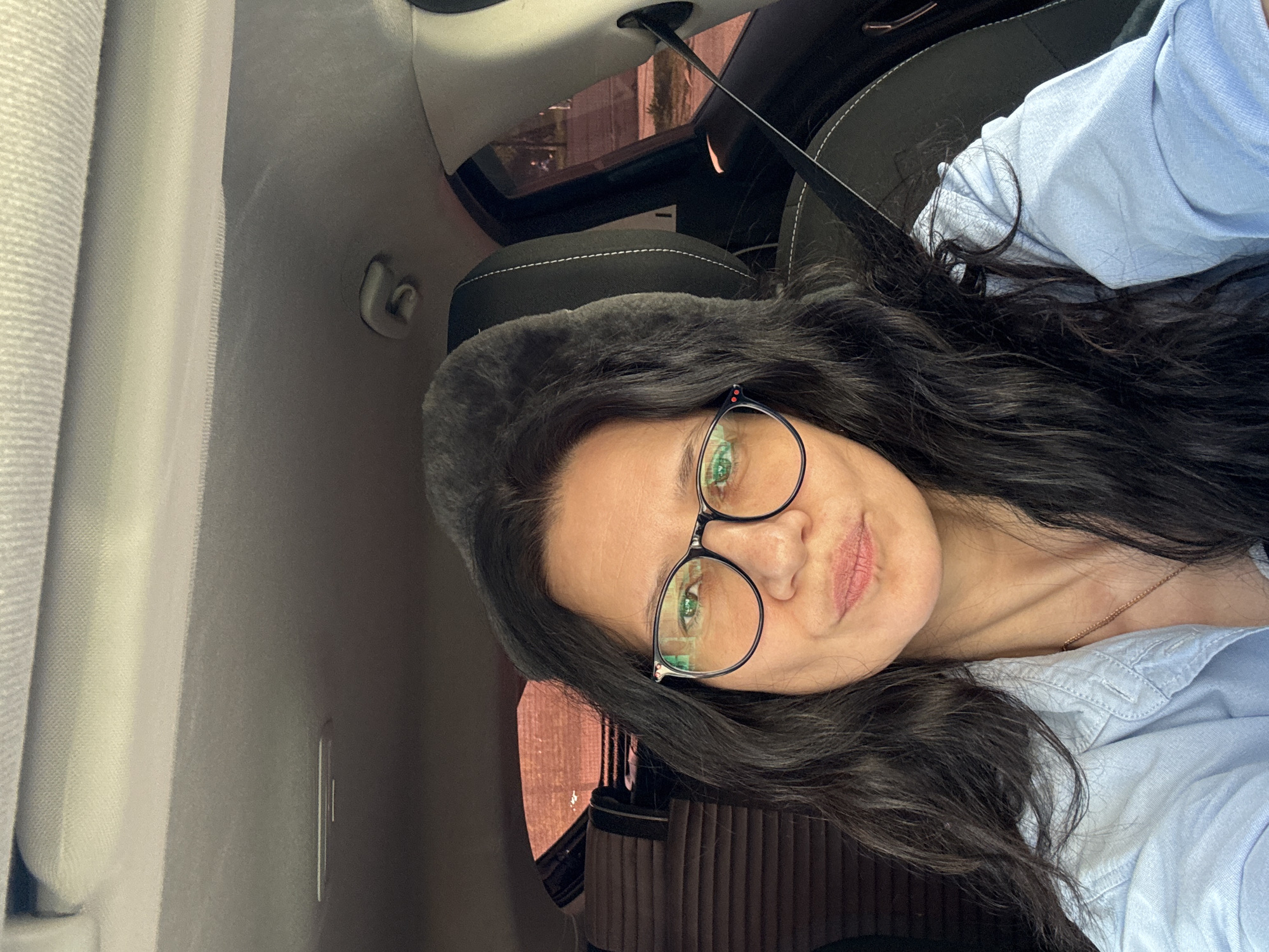 A woman with dark hair and glasses is in a car. She is wearing a light blue shirt and a necklace. The car has black seats and a sunroof. The image was likely taken in Chaikovsky, Russia, although this cannot be definitively determined from the image alone.