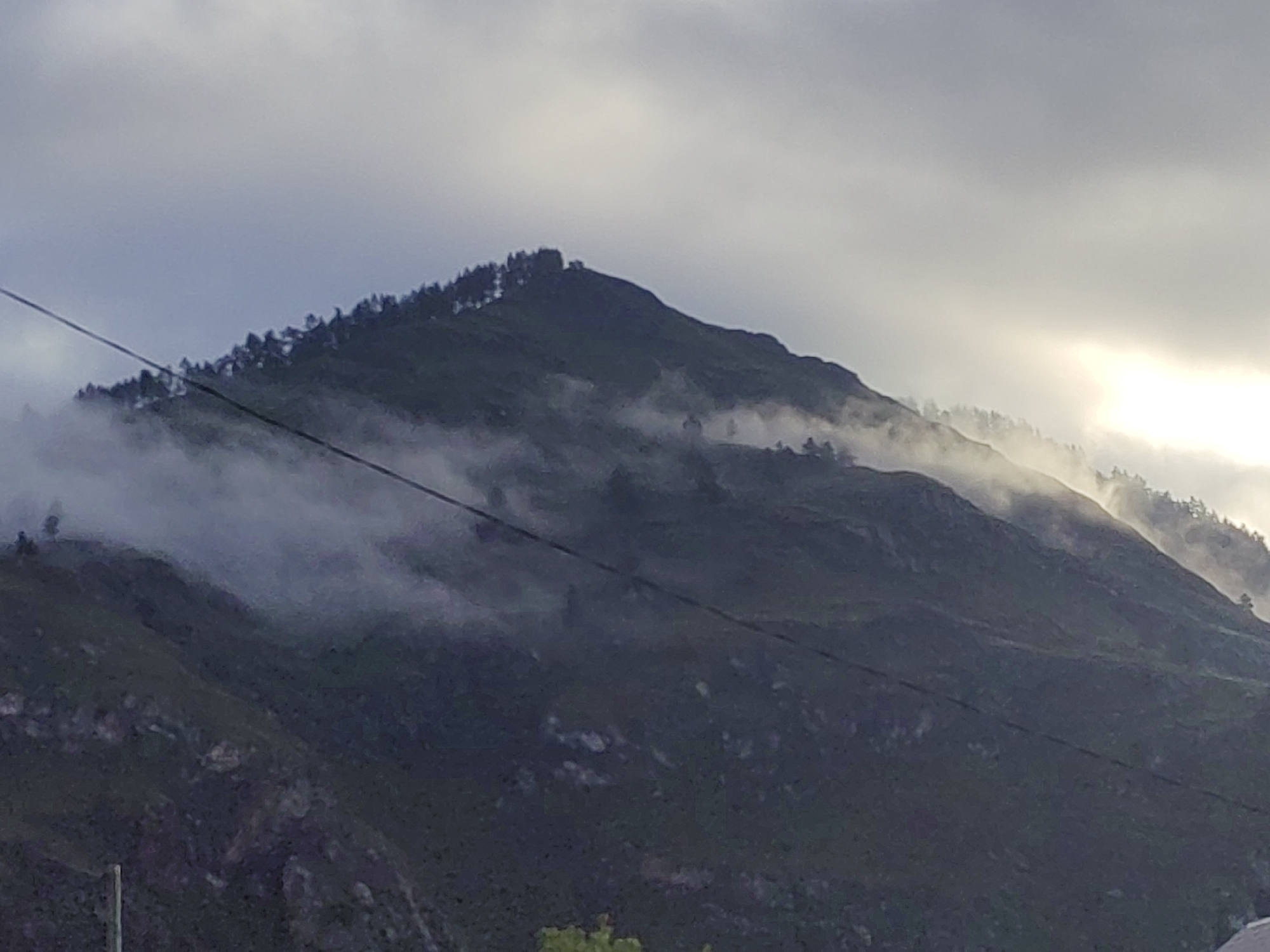 The image shows a dark, misty mountain range in Anos, Russia. The peak of the mountain is covered in trees, and wisps of fog snake around its slopes. A wire runs diagonally across the frame, obscuring parts of the mountain and sky. The sky above is a mix of grey clouds and bright, hazy sunlight peeking through. The overall mood is somber and mysterious, with the fog and dark tones contributing to a sense of isolation.