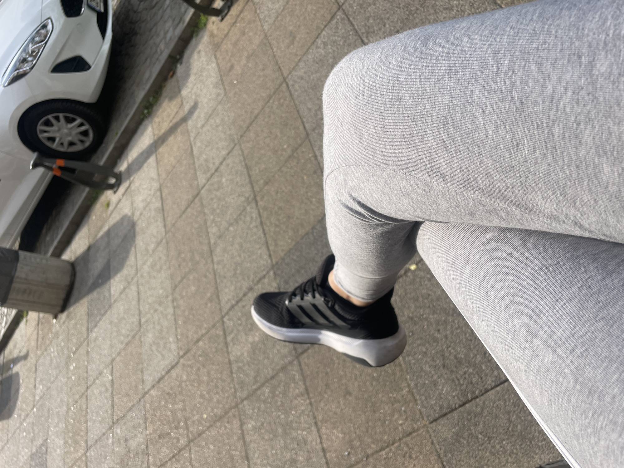 A person's legs, clad in light gray leggings and black athletic shoes with white soles and accents, are the primary focus of the image. The legs are crossed, with the right leg positioned over the left. The shoes display the distinctive three stripes of the Adidas brand. The scene appears to be outdoors on a sidewalk paved with square, light-colored tiles. In the background, a white car is parked, and a dark gray cylindrical trash can is visible on the left. The lighting suggests it is daytime, with shadows cast on the pavement, indicating sunlight. The specific location in Stara Zagora, Bulgaria cannot be determined from the visual information provided.