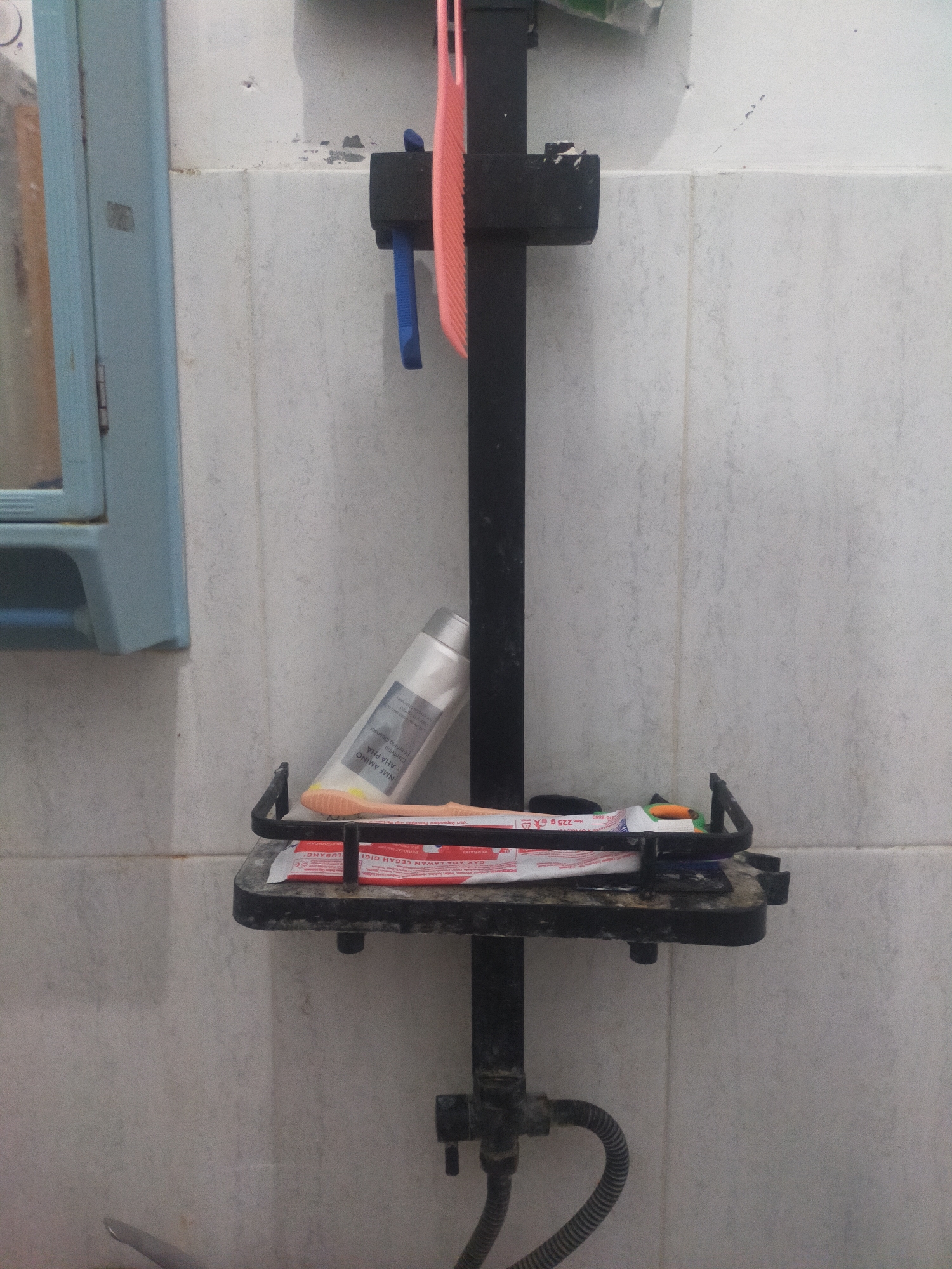 The image shows a bathroom shelf with toiletries. A black metal pole supports a shelf holding a tube of toothpaste, toothbrushes, and a comb. The wall behind the shelf is tiled with white squares. To the left, a portion of a light blue medicine cabinet is visible. A flexible grey tube connects to the bottom of the pole. The scene appears to be in a residential bathroom, likely in Tegalandong, Indonesia, given the presence of the Indonesian text on the toothpaste packaging. There are no people or actions depicted in the image.