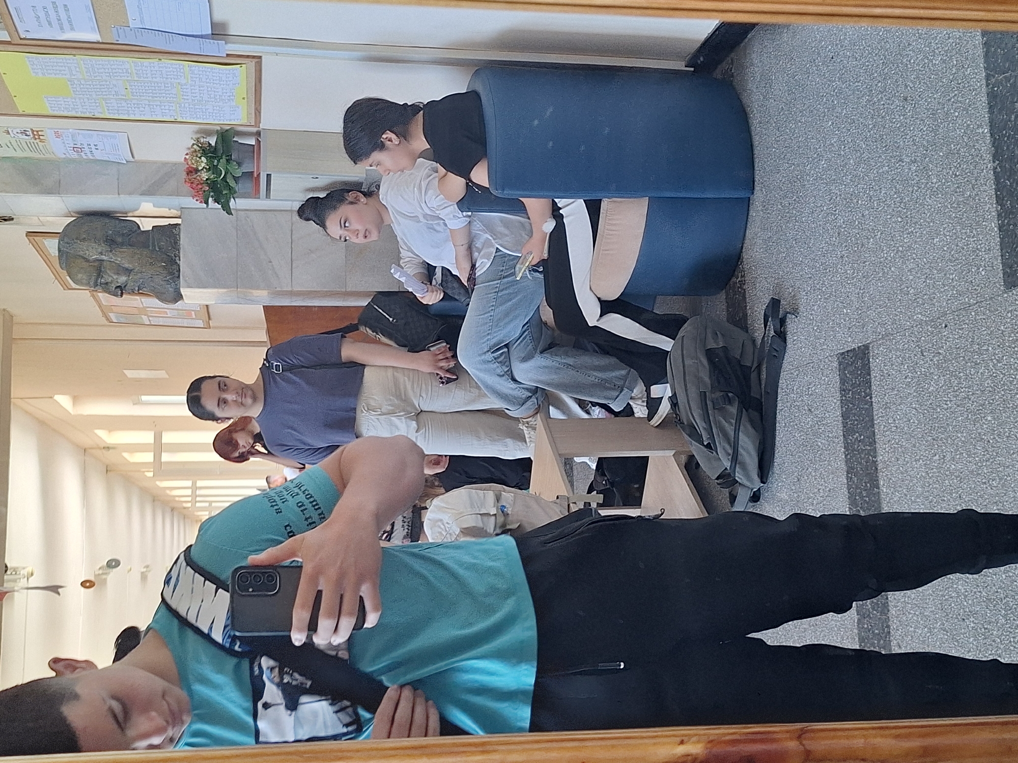 The image depicts a reflection in a mirror of a hallway in what appears to be a school or university building in Varna, Bulgaria. In the foreground, a young person, seen from the chest down, is taking a selfie. They are wearing a teal t-shirt, black pants, and a black backpack with a white and blue strap.
In the background, reflected in the mirror, several other individuals are visible. Two young women are seated in a blue armchair. The woman on the left is wearing a white long-sleeved shirt and light blue jeans, holding a piece of paper and a phone. The woman on the right is wearing a black t-shirt and black pants with a white stripe down the side, also holding a phone. Another young woman with red hair is standing behind them, looking down. Further down the hallway, another person is partially visible.
The hallway features light-colored walls with bulletin boards displaying various notices and charts. A bust of a man is mounted on the wall. Potted plants with pink and orange flowers are placed near the bust. The floor is a speckled grey material with dark grey rectangular patterns. A wooden desk or table is positioned in the middle of the hallway. A grey backpack lies on the floor near the seated women. The overall impression is of a casual gathering within an academic setting.