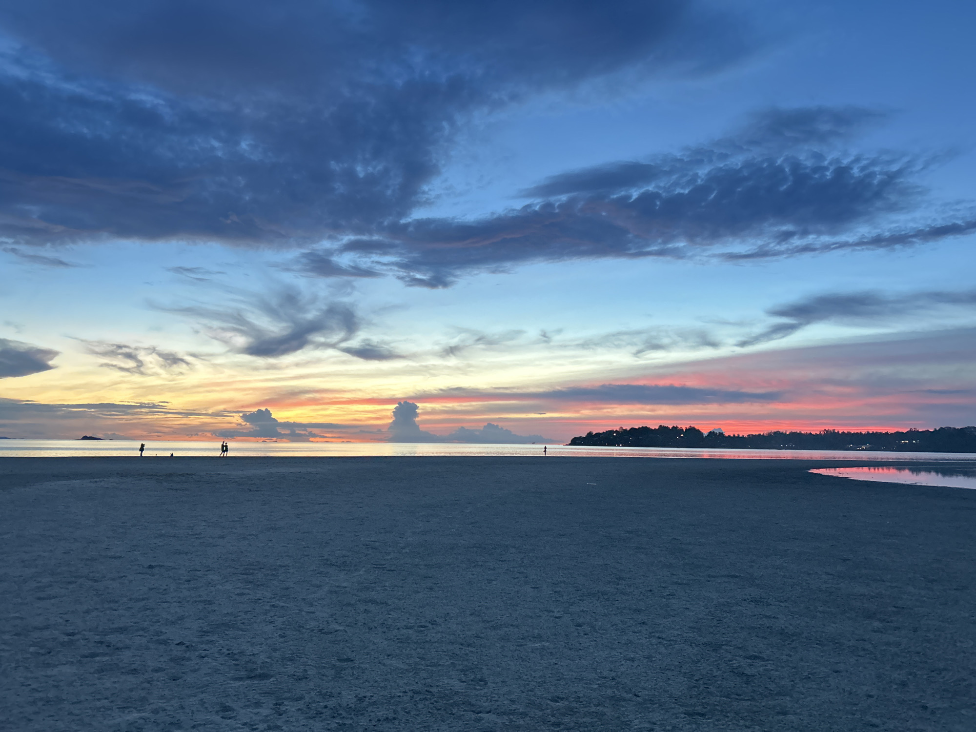The image depicts a serene beach scene at dusk in the village of Koh Phangan, Thailand. The sky is a vibrant canvas of blues, pinks, and oranges, with dramatic clouds casting shadows. The foreground is dominated by a wide expanse of dark, sandy beach, with subtle textures suggesting fine grains. In the distance, the calm ocean reflects the colors of the sunset. A silhouetted treeline marks the horizon, with a few scattered lights indicating buildings. Three small human figures are visible on the beach, one standing alone, and two others in close proximity, possibly engaged in conversation or walking together. Another solitary figure is visible standing near the water's edge further down the beach. The overall impression is one of tranquility and natural beauty.