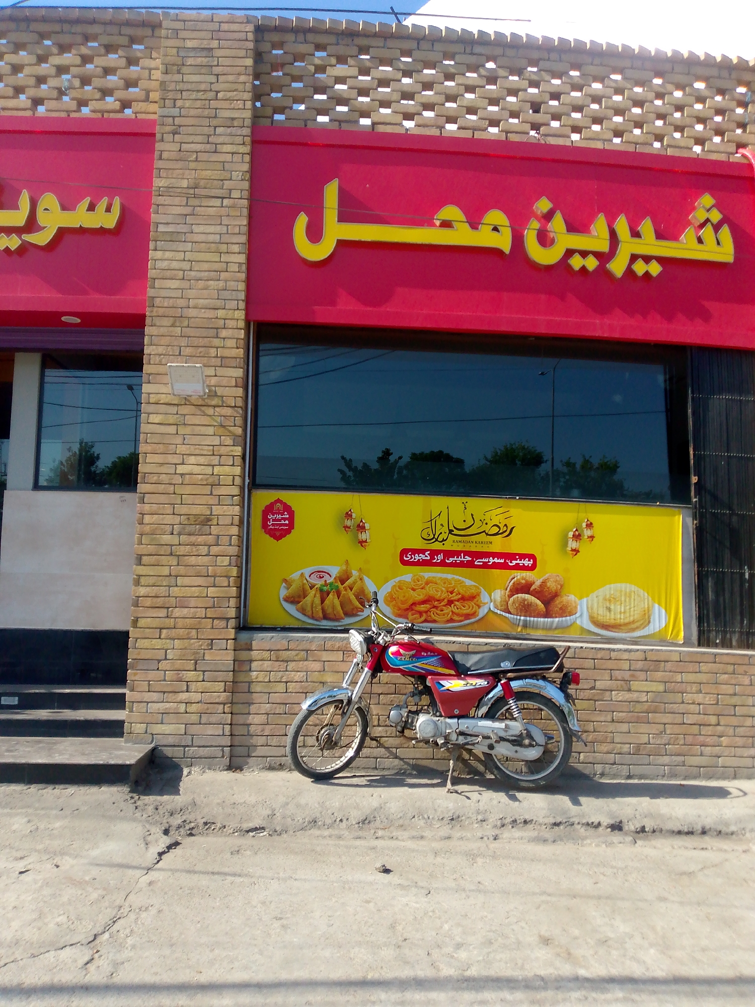 The image shows the exterior of a shop with red and yellow signage. The shop has a brick facade and a large glass window. A red motorcycle is parked on the sidewalk in front of the shop. A yellow banner is displayed in the window, featuring images of food items and text in an Urdu script. The text in the banner appears to be a Ramadan greeting, with "Ramadan Kareem" written in English as well. The sign above the window reads "شیرین محل" in yellow Urdu script on a red background. Another sign to the left, partially visible, also has yellow Urdu script on a red background. The setting appears to be a street in an urban environment. The lighting suggests it is daytime, with shadows cast by the motorcycle and the building.