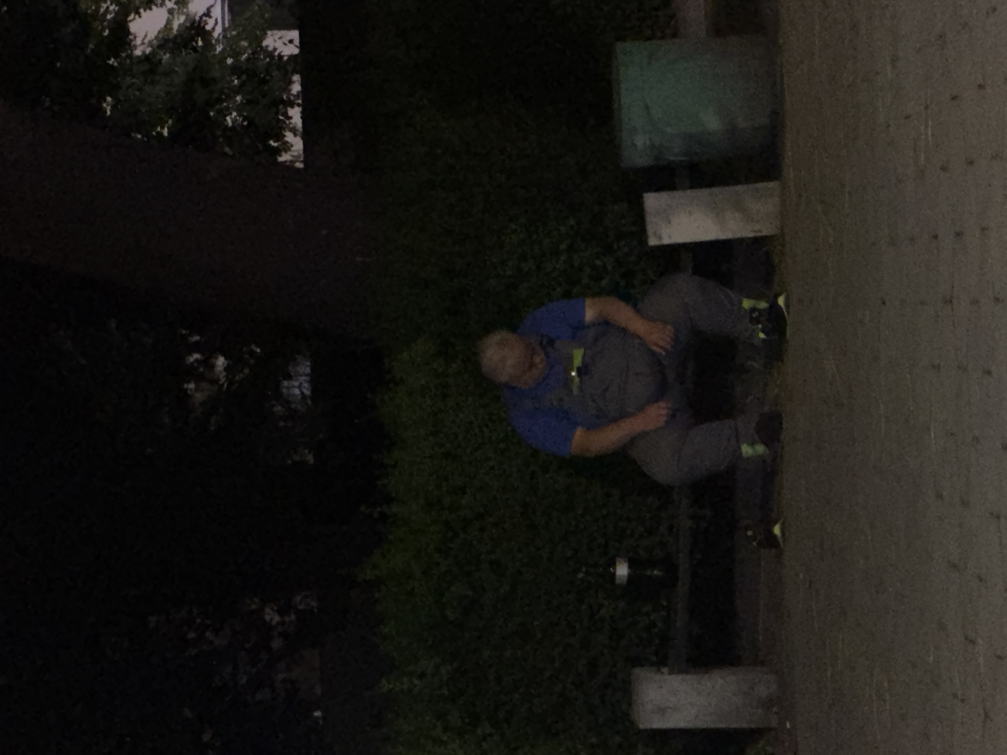 A man in a blue shirt and gray overalls is sitting on a bench, slumped over with his head down. He is wearing black shoes with yellow accents. Next to him on the bench is a bottle. To his left is a concrete block, and to his right are two more concrete blocks, one white and one green. The bench is in front of a dense green hedge. The scene is dimly lit, suggesting it is nighttime or very early morning. The image appears to be taken in an outdoor urban setting, possibly a park or public space in Plovdiv, Bulgaria.