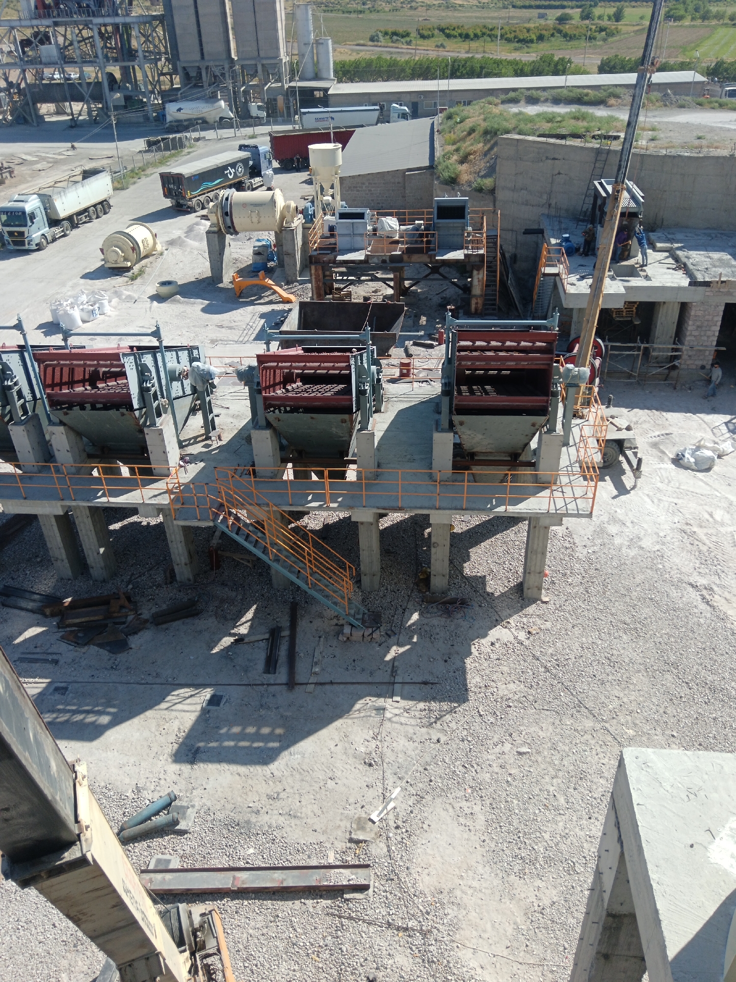 This is a high-angle, exterior, medium shot of a construction site in Aragatsavan, Armenia. The scene depicts an industrial area with heavy machinery, including crushers, screens, trucks, and a crane. In the foreground, there are two large, multi-deck screen machines supported by concrete pillars. Orange railings and stairs provide access to these elevated platforms. In the middle ground, several trucks are present, including a white tanker truck, a blue semi-trailer truck, and a red dump truck. A large concrete batching plant with silos is visible in the background. Several construction workers are seen working on the site, with one appearing to be operating the crane. The ground is covered in gravel and dust, indicative of an active construction environment. The surrounding landscape features dry vegetation and low-lying hills under a clear sky.
