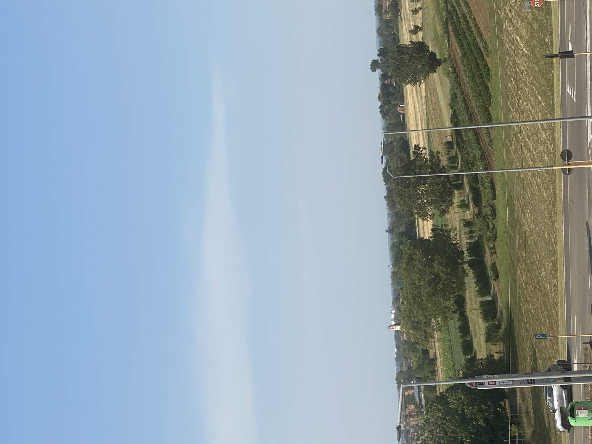 The image captures a rural landscape near Parma, Italy, under a clear blue sky. In the foreground, a road is visible, with a silver car parked to the left, next to a green bin. A bus stop sign stands prominently on the left side of the road. Beyond the road, a field stretches out, featuring rows of crops, interspersed with patches of dry grass and trees. A tall, slender church steeple rises in the distance, suggesting a nearby settlement. The overall scene depicts a typical rural vista, with agricultural land and scattered buildings.