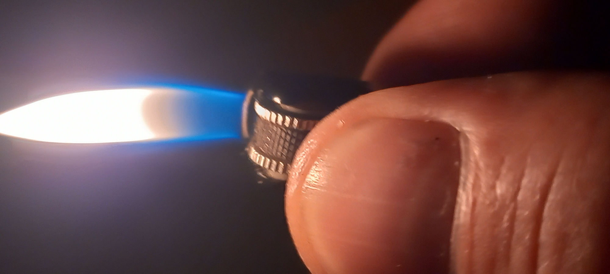 The image shows a close-up of a person's thumb holding a lit lighter. The flame is bright white at the base with a blue halo, transitioning to a yellowish-orange at the top. The lighter has a ridged metal wheel for ignition. The thumb appears to be a left thumb due to the position of the cuticle and nail. The background is dark and out of focus. There is no identifiable information about the location in Kryvyi Rih, Ukraine.