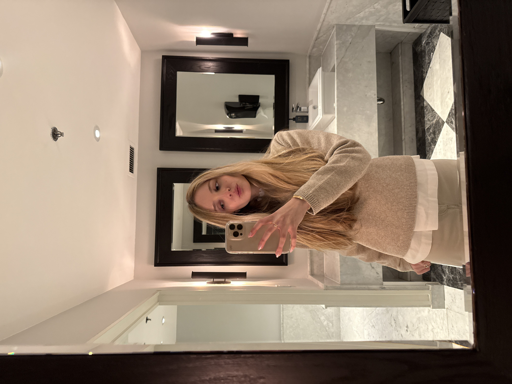A young woman takes a selfie in a bathroom, visible in the mirror. She has long blonde hair and wears a beige sweater over a white shirt. She holds a beige smartphone up to her face with her right hand, displaying a gold ring on her finger.
Behind her, the bathroom features two large, dark-framed square mirrors above a white sink and marble-patterned wall tiles. A black object resembling a toilet paper dispenser is visible in the top mirror. The bathroom is lit by overhead lights and wall sconces.
It's not possible to determine the location in Mexico City, Mexico, based on the image alone.