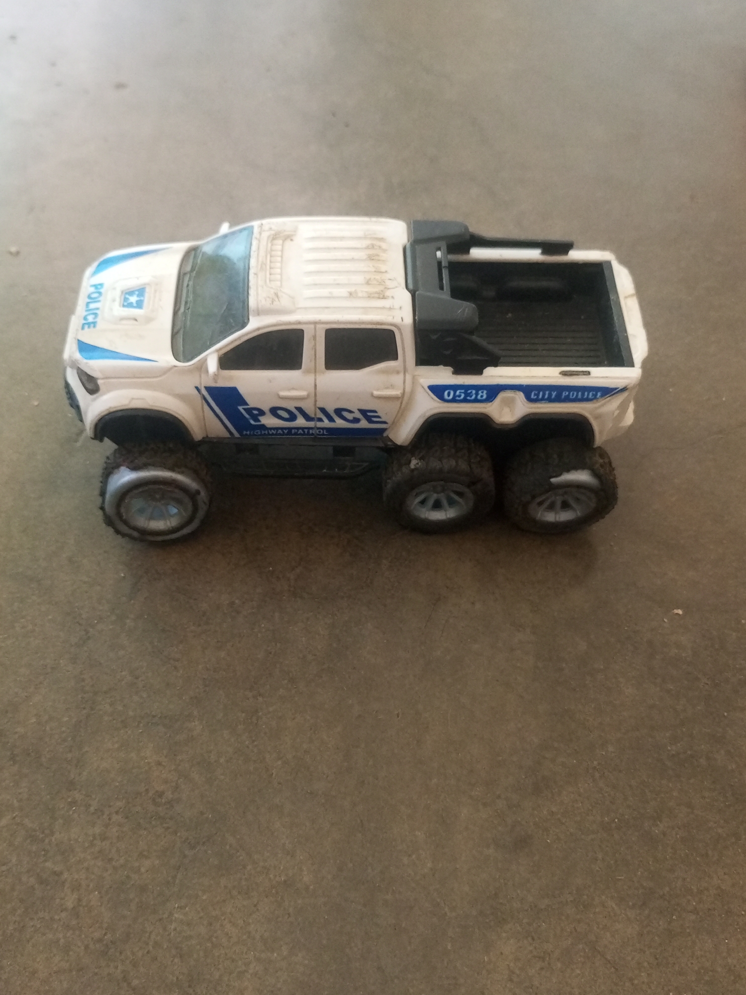 A toy police truck, likely for children, rests on a concrete surface. The truck is white with blue accents and features the word "POLICE" prominently displayed. It has six large, rugged tires and a six-wheeled drive system. The truck appears to be a model of a pickup truck with an open bed in the rear. The concrete surface suggests an outdoor setting, possibly a sidewalk or road. There are no people or animals visible in the image, nor any indication of a specific location within La Tagua, Venezuela.