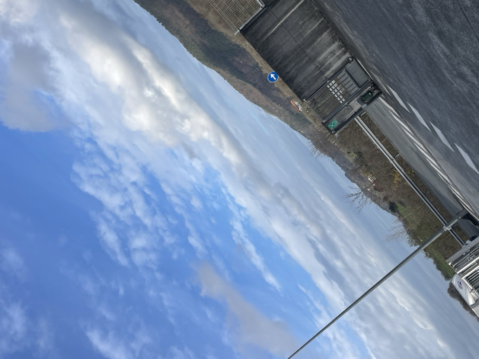 This image, captured in the city of Oiartzun, Spain, presents an outdoor scene under a partly cloudy sky. The photograph is tilted, aligning the horizon diagonally from the lower left to the upper right. The sky, occupying the upper left two-thirds of the frame, is bright blue with scattered white and light grey clouds. In the background on the right, a verdant mountain range is visible, covered in trees, some appearing deciduous with sparse foliage, suggesting late autumn or winter. A single structure with a red or orange roof is discernible on a distant hillside.
In the mid-ground on the right, a grey concrete wall topped with a metal fence extends horizontally. A blue circular road sign, displaying a white upward-pointing arrow, is affixed to the wall. Adjacent to it is a green square sign with white symbols. Below the wall, a dark metal gate is present. The foreground features a section of asphalt road with white chevron-style road markings. A grey metal guardrail separates the road from the hillside. A tall, slender grey street lamp post rises from the lower left portion of the frame, extending towards the center-top. A partial view of a light-colored vehicle, possibly the front section of a truck or trailer, is visible in the bottom-left corner. No individuals are present in the scene, and no explicit actions are depicted.