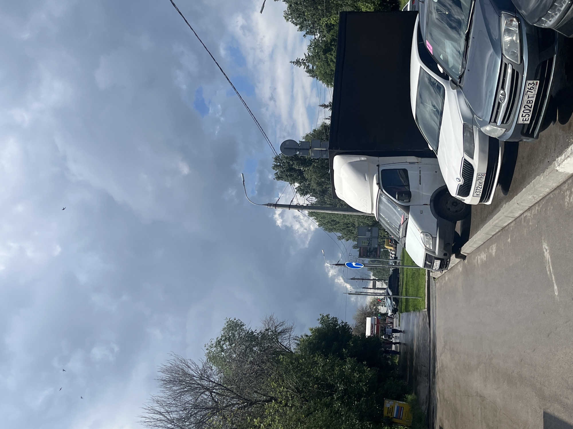 In an outdoor, urban environment under a cloudy sky, several vehicles are parked along a street. In the foreground, a dark gray sedan with the license plate E502BT763 is parked, facing left. To its right and slightly behind it, a white car with the license plate X372XK763 is also parked. Further back, a white van with a black cargo box is visible. The street appears wet, suggesting recent rain. Trees line the left side of the road, and a few pedestrians are walking in the distance on the sidewalk. There are streetlights and traffic signs visible. Several birds are flying in the sky.