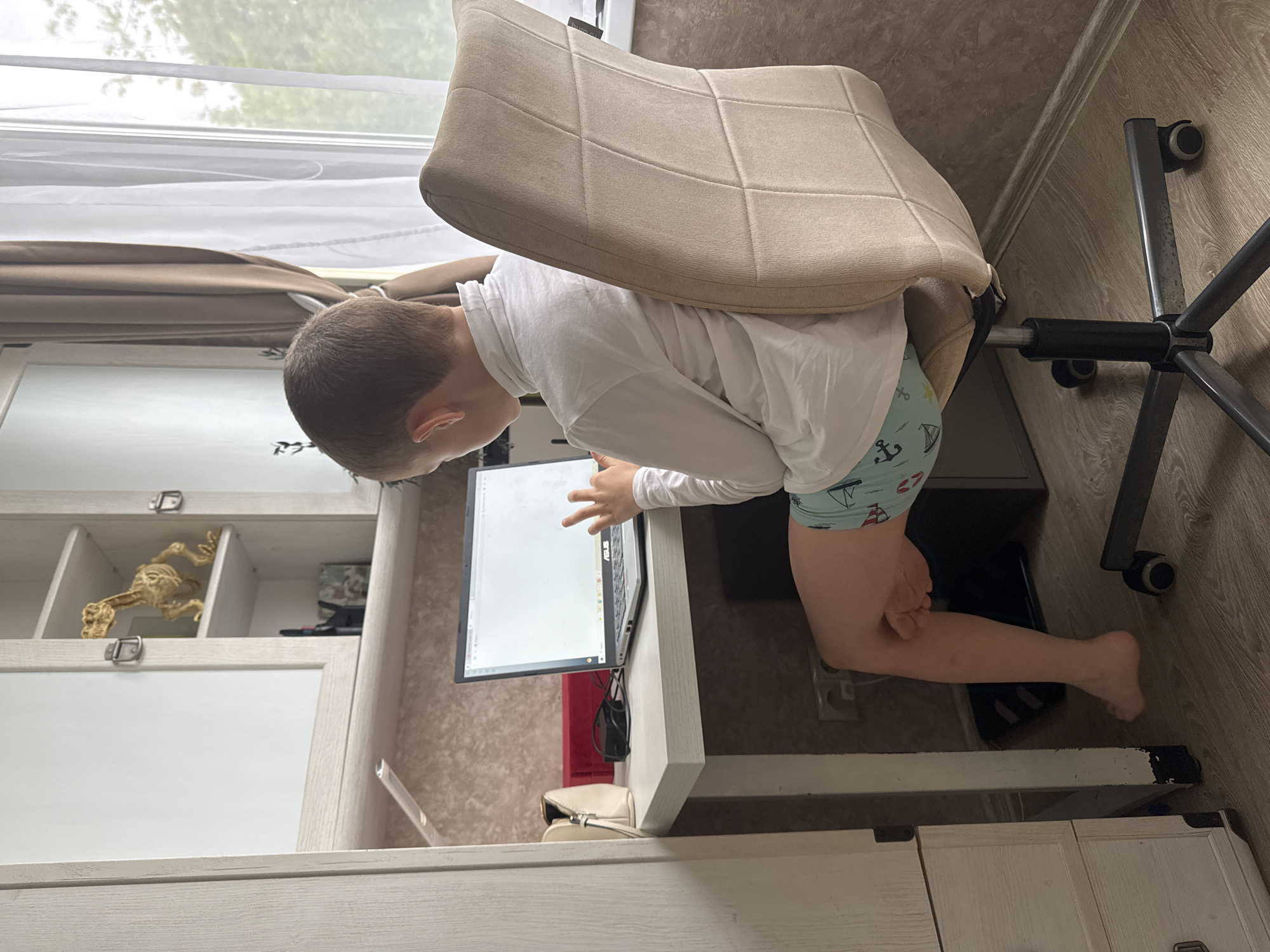 A child in a white long-sleeved shirt and patterned underwear is leaning over a desk looking at a laptop screen. The child is in a room with a window, a wardrobe with a decorative skeleton on a shelf, and a rolling office chair. The scene appears to be indoors, with the furniture and objects suggesting a domestic environment. There is no explicit evidence to confirm the location as Раздолье, Russia; this would require additional information not present in the image.