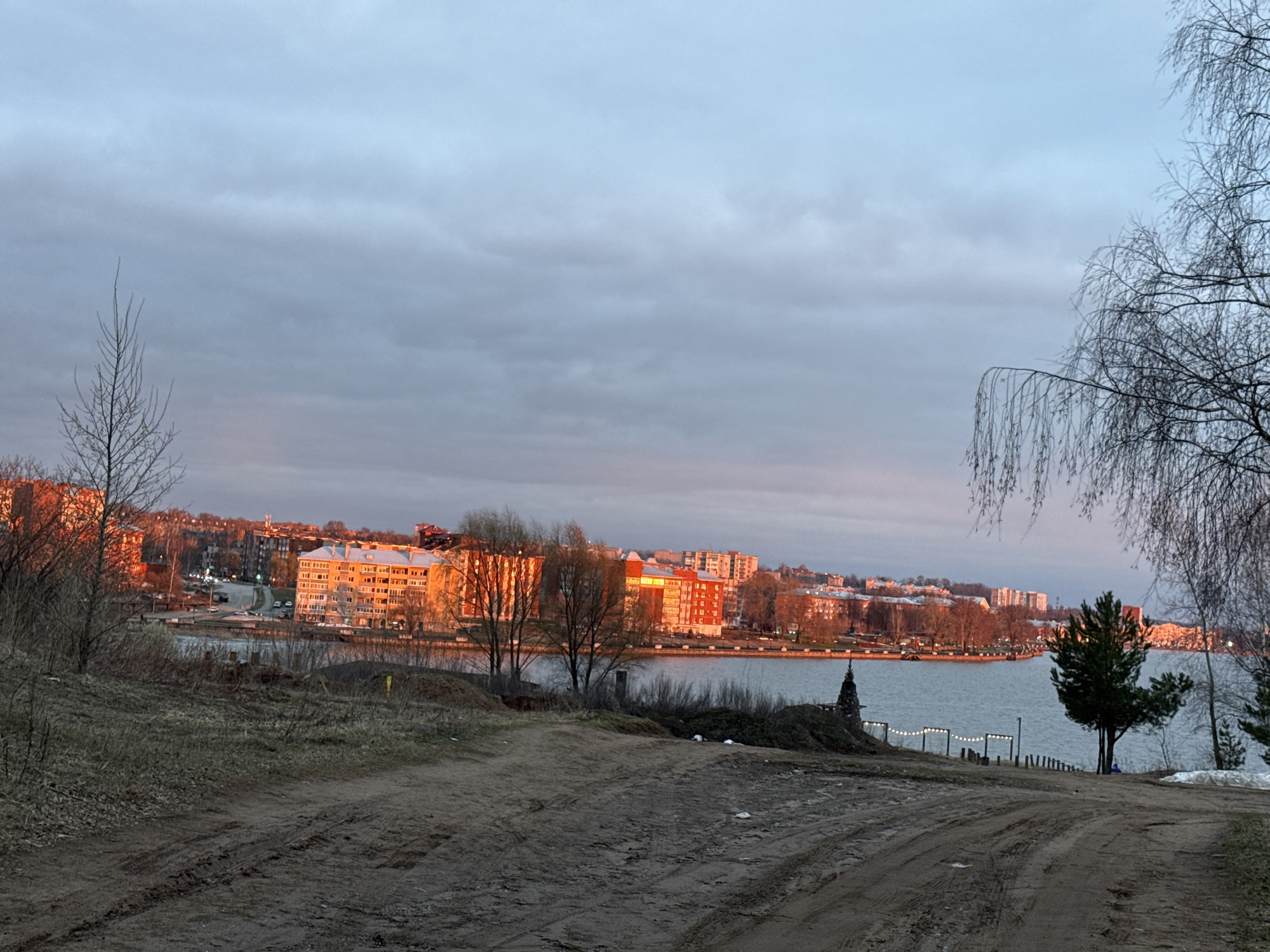 The image presents a wide landscape view of a town or city, likely Votkinsk, Russia, situated along a large body of water, possibly a lake or reservoir. In the foreground, a prominent unpaved dirt path, marked with tire tracks, slopes gently downwards from the left towards the center and right. The ground is covered with sparse, dry vegetation and some exposed soil, suggesting a late autumn, winter, or early spring season.
Across the water, on the opposite bank, a line of multi-story buildings, predominantly residential or mixed-use, catches the warm, golden-orange light of a low sun. This light brightly illuminates their facades, creating a striking contrast with the overcast sky and the muted tones of the foreground. Other buildings and distant urban development are visible further back on the horizon. The water itself appears calm and reflective.
Trees are a significant element in the composition. A slender, bare deciduous tree stands on the left against the sky, while on the right, the graceful, drooping branches of a larger, bare tree (resembling a weeping birch) frame the scene. Closer to the water's edge on the right, a small evergreen tree stands, with a patch of melting snow nearby. A string of decorative white lights is visible along the shoreline on the right, strung between posts, suggesting a promenade or public space. A small, dark, roughly conical structure is also present near the water on the right.
The sky is largely overcast with soft, diffuse clouds, displaying shades of light grey and blue. The warm illumination of the distant buildings indicates either sunrise or sunset. The absence of foliage on most trees further points to a cool season. No people are visible, and there is no discernible text in the image. The scene conveys a tranquil, slightly melancholic atmosphere.