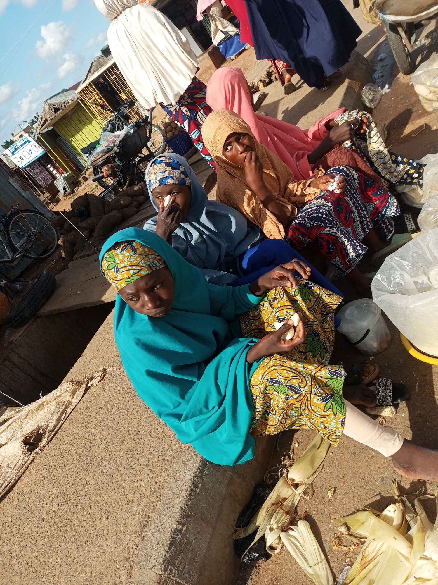 In Tempure, Nigeria, three women in vibrant hijabs are seated outdoors, possibly at a market. The woman in the foreground wears a turquoise hijab and a yellow and green patterned dress, holding what appears to be food in her hands. To her right, another woman, also in a patterned dress, wears a beige hijab and is seen touching her face. Behind them, a third woman in a blue hijab is visible. Their clothing suggests a warm climate. The environment appears to be a dusty, outdoor setting, with debris including corn husks scattered on the ground. In the background, there are market stalls and a bicycle, indicating a busy marketplace. The lighting suggests it is daytime, with sunlight casting shadows.
