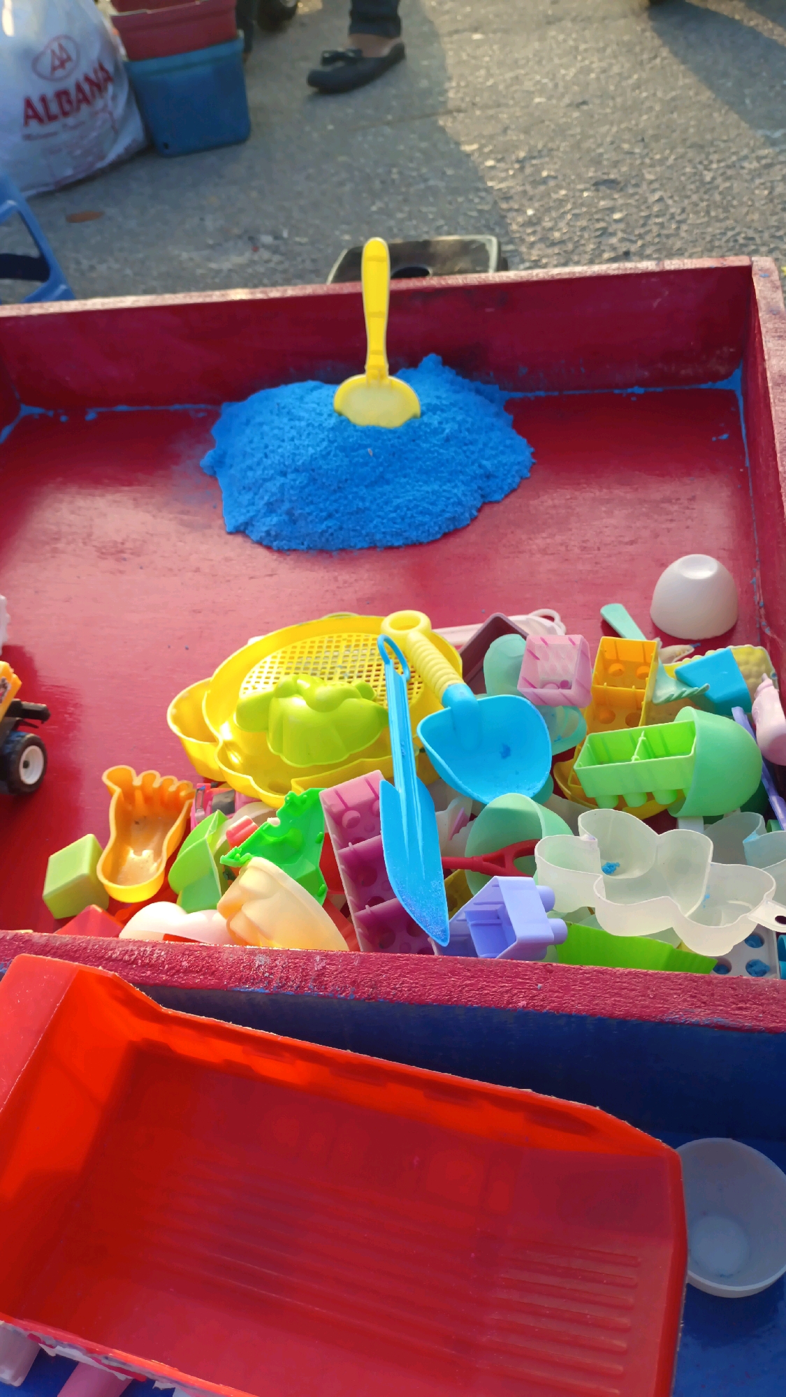 An outdoor scene depicts a red sandbox filled with various colorful plastic toys for children. In the center of the sandbox, a pile of bright blue kinetic sand is visible, with a small yellow plastic shovel embedded in it. Scattered throughout the sandbox are numerous other plastic toys, including sieves, molds, buckets, and shovels in shades of yellow, green, blue, purple, and white. In the foreground, a large red plastic container or tray is partially visible, also containing some toys. The ground surrounding the sandbox appears to be paved asphalt, with a shadow cast by an unseen object. In the background, the legs of a person wearing dark pants and dark shoes are visible, suggesting an adult is present. A white plastic bag with the "ALBANA" logo and some red and blue buckets are also visible in the background. The scene appears to be set up for children's play, possibly at a park or a market stall.