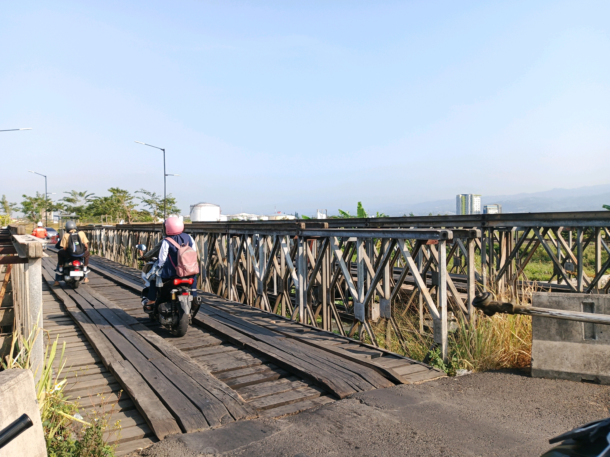 The image depicts a perspective view of a wooden bridge on a sunny day. In the foreground, two individuals on motorcycles are traversing the bridge. The first motorcycle, closer to the viewer, carries a rider with a pink helmet and a pink backpack. The second motorcycle, slightly ahead and to the left, has a rider wearing a helmet and a yellow backpack. The bridge itself is constructed with weathered wooden planks laid across a steel truss structure. Beyond the bridge, the landscape features vegetation, industrial facilities with large white storage tanks, and distant buildings, with a clear blue sky overhead. The overall scene suggests a functional, somewhat rustic, infrastructure within an urban or semi-urban environment. The location is identified as Bandung City, Indonesia.