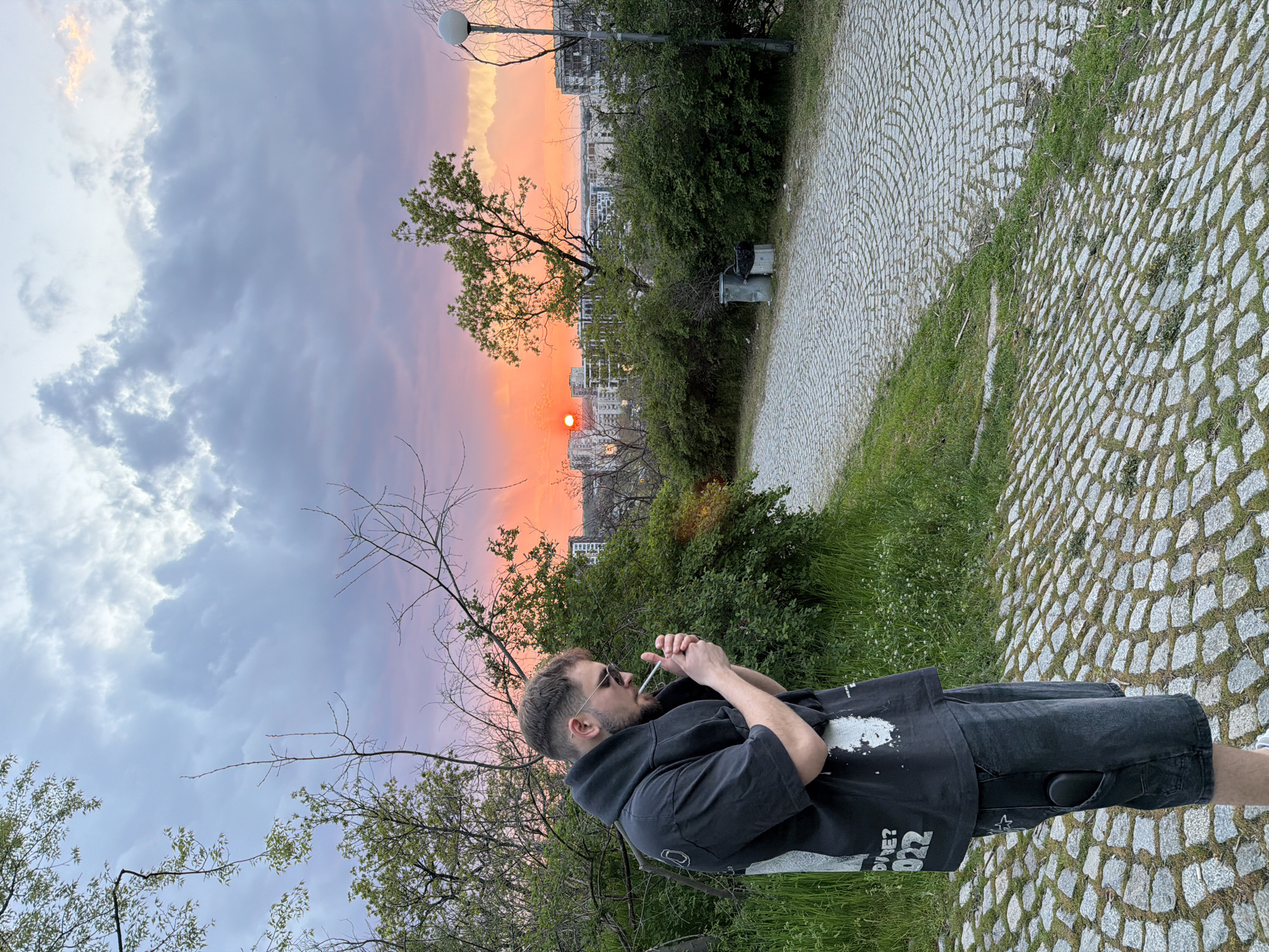 A man is standing on a cobblestone surface near vegetation, holding a cigarette in his mouth with his hands crossed. He wears a black hooded sweatshirt and dark shorts. The background features a sunset, trees, and buildings, a street light. The cobblestone extends into the distance, and the scene appears to be at dusk or dawn. The photo appears to be taken at the top of Bunardzhik Hill in Plovdiv, Bulgaria.