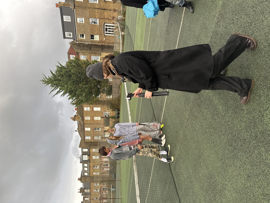 A group of people are standing on a tennis court. A person in a black coat and beanie holds a phone on a tripod, possibly filming the others. Three people stand together, holding tennis rackets. One wears camo pants, another a pink flannel, and the third a gray shirt. Behind them, there is a tennis net and a row of residential buildings under a cloudy sky. A tall evergreen tree is also visible near the buildings.