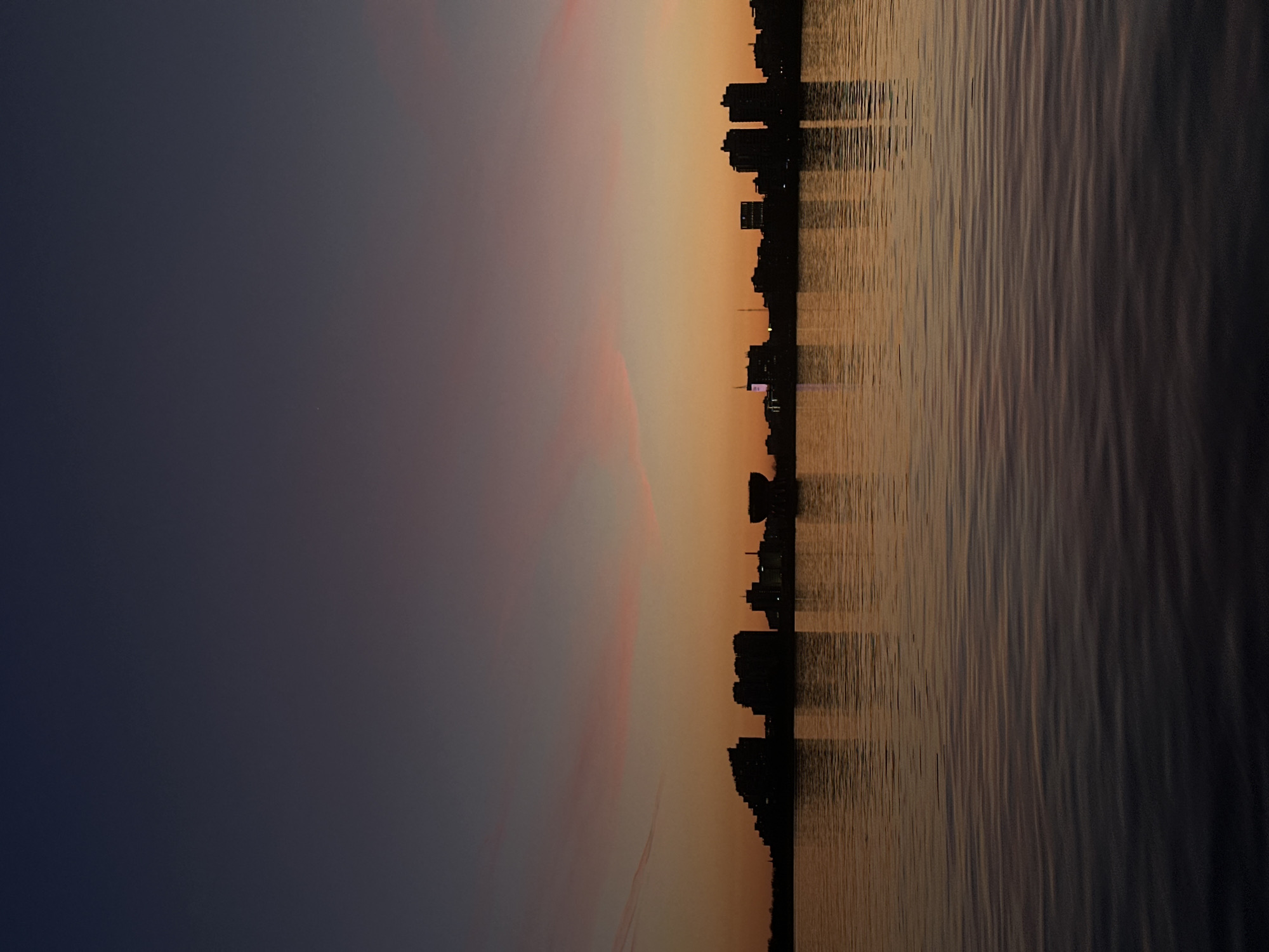 The image captures a serene twilight scene of a city skyline across a body of water, likely in Kazan, Russia. The sky dominates the upper part of the frame, transitioning from a deep, dark blue-purple at the top to a warm gradient of orange, yellow, and subtle pink hues closer to the horizon, indicative of a sunset or sunrise. Faint, diffuse cloud formations are visible within the lighter sections of the sky.
Along the horizontal mid-line, a dark silhouette of a city skyline stretches across, featuring numerous buildings of varying heights, including multi-story structures and possibly some with distinct architectural elements. A few small, distant lights can be seen twinkling within the dark mass of the buildings.
The lower half of the image is comprised of a calm body of water, reflecting the vibrant colors of the sky. The water's surface shows gentle ripples and texture, clearly mirroring the golden and orange tones from the horizon. The reflections of the city buildings appear as elongated dark streaks extending downwards into the water.
The overall atmosphere is peaceful and contemplative, highlighting the tranquil beauty of the transitional time of day. There are no people or discernible text visible in the image. The weather appears clear and still.