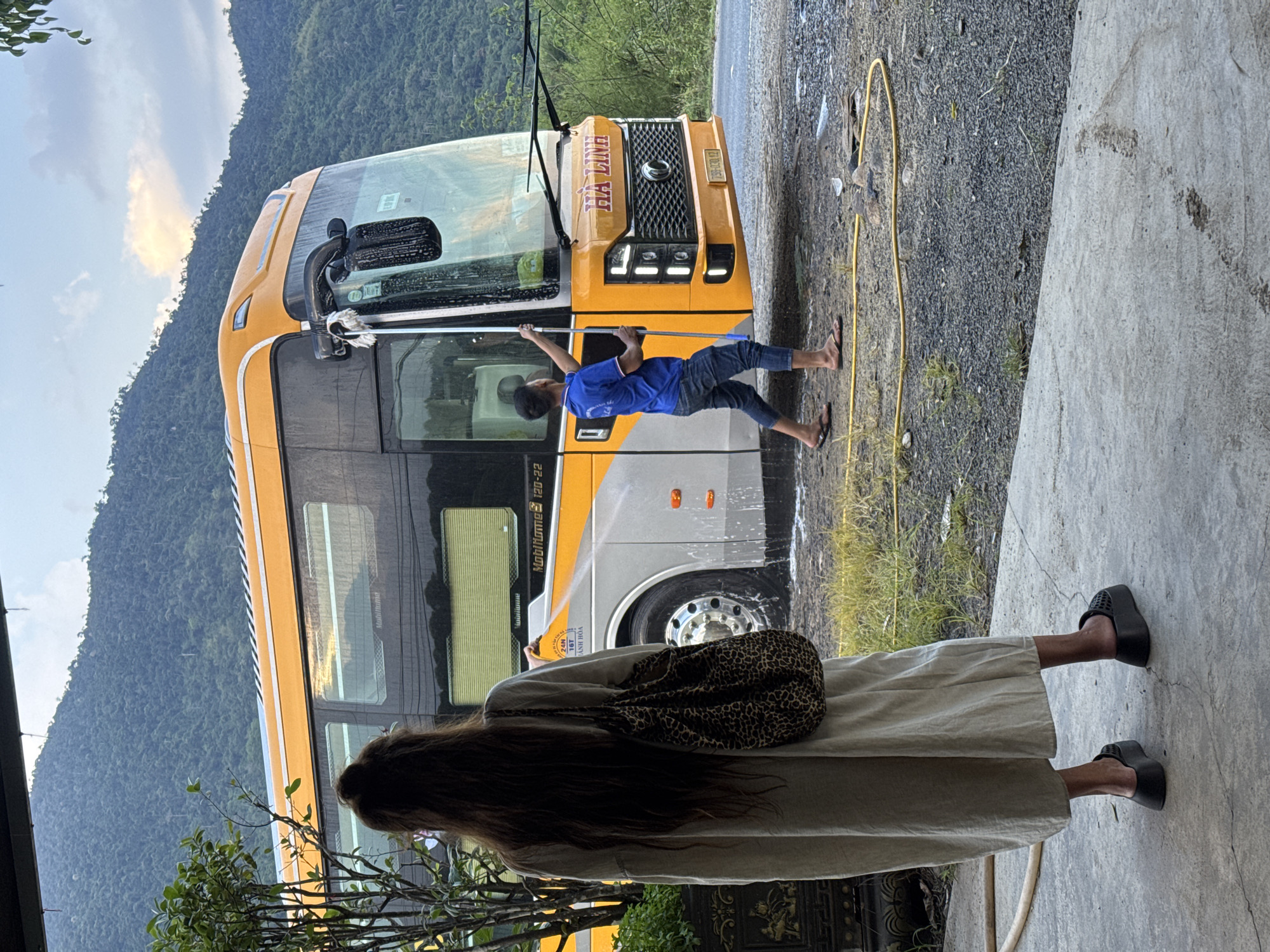A person is washing a yellow bus next to a building in Tây Ninh Hòa, Vietnam. The bus has the words "Hà Linh" on the front. A person with long hair, wearing a tan dress and black sandals, stands near the bus. The surrounding environment features mountains, and the ground is a mix of concrete and gravel.