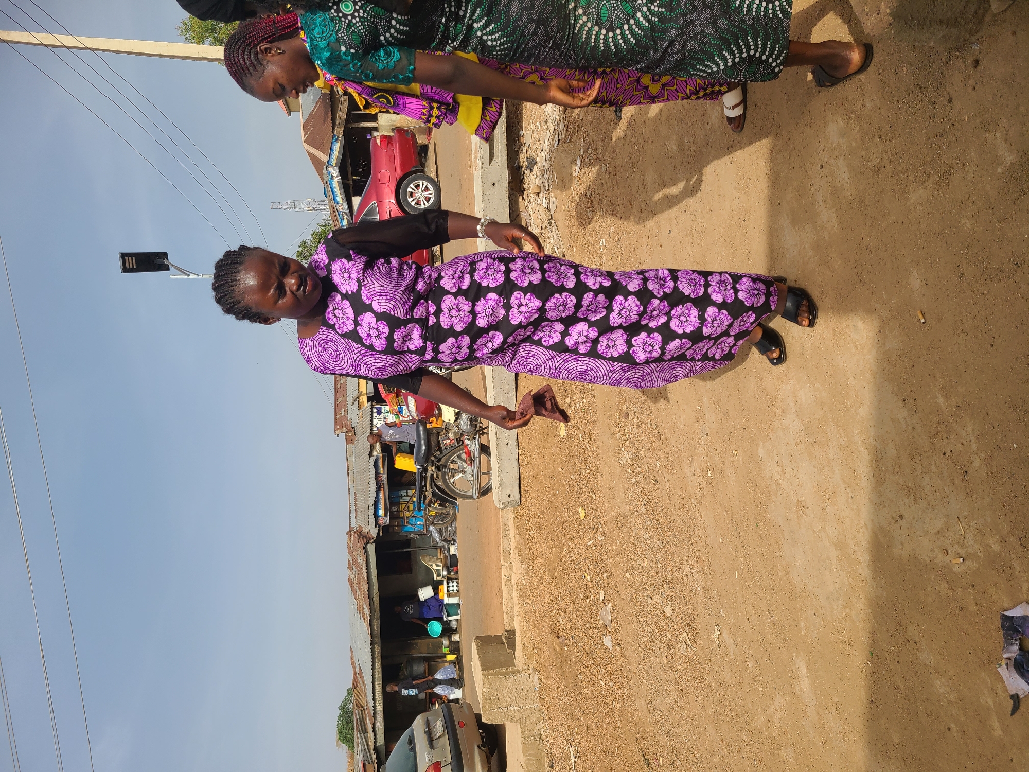 Two women are standing on a dirt road next to an urban street. The woman in the foreground is wearing a long purple and black flower print dress. The other woman, closer to the upper edge, is in a long green and black print dress with a pink and yellow layer. They are both wearing flat sandals. The background shows a typical African street scene with a red car, motorcycles, and small shops or buildings with corrugated metal roofs. A utility pole and power lines are visible in the sky. The picture is shot from a low angle and rotated 90 degrees clockwise. The location is implied to be Jos, Nigeria based on the prompt.