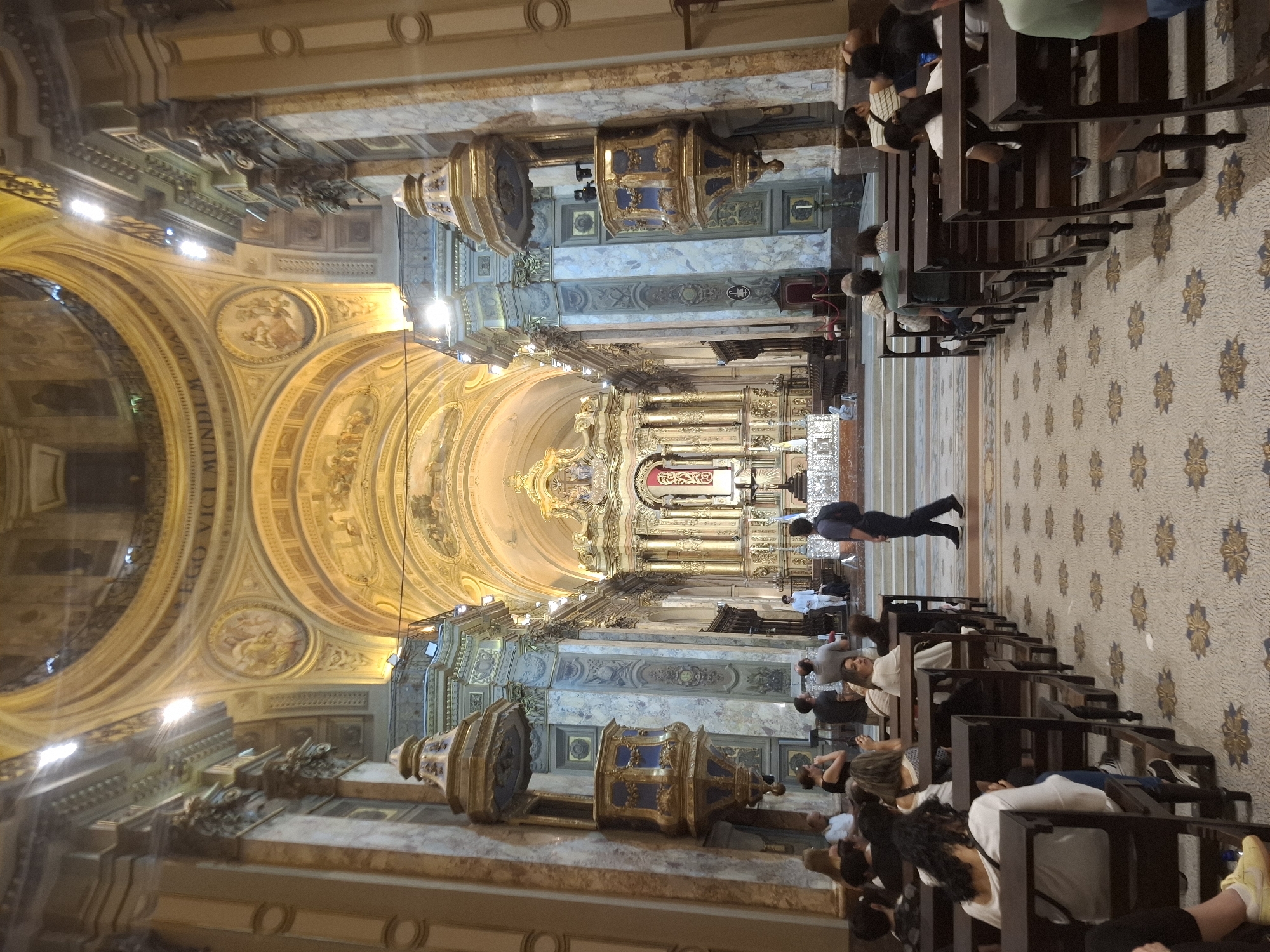 Captured within the architectural grandeur of the Buenos Aires Metropolitan Cathedral in Argentina, the image offers an interior perspective of the basilica. Elaborate domed ceilings are adorned with ornate artwork, cascading toward the high altar at the far end of the hall. The nave's floor, patterned in mosaic tiles, stretches from the foreground to the altar. Wooden pews line the aisle, occupied by a congregation of people attending a service or visiting. A man walks along the aisle. Lecterns are positioned at intervals along the walls. The scene conveys a sense of reverence within a historic religious setting.