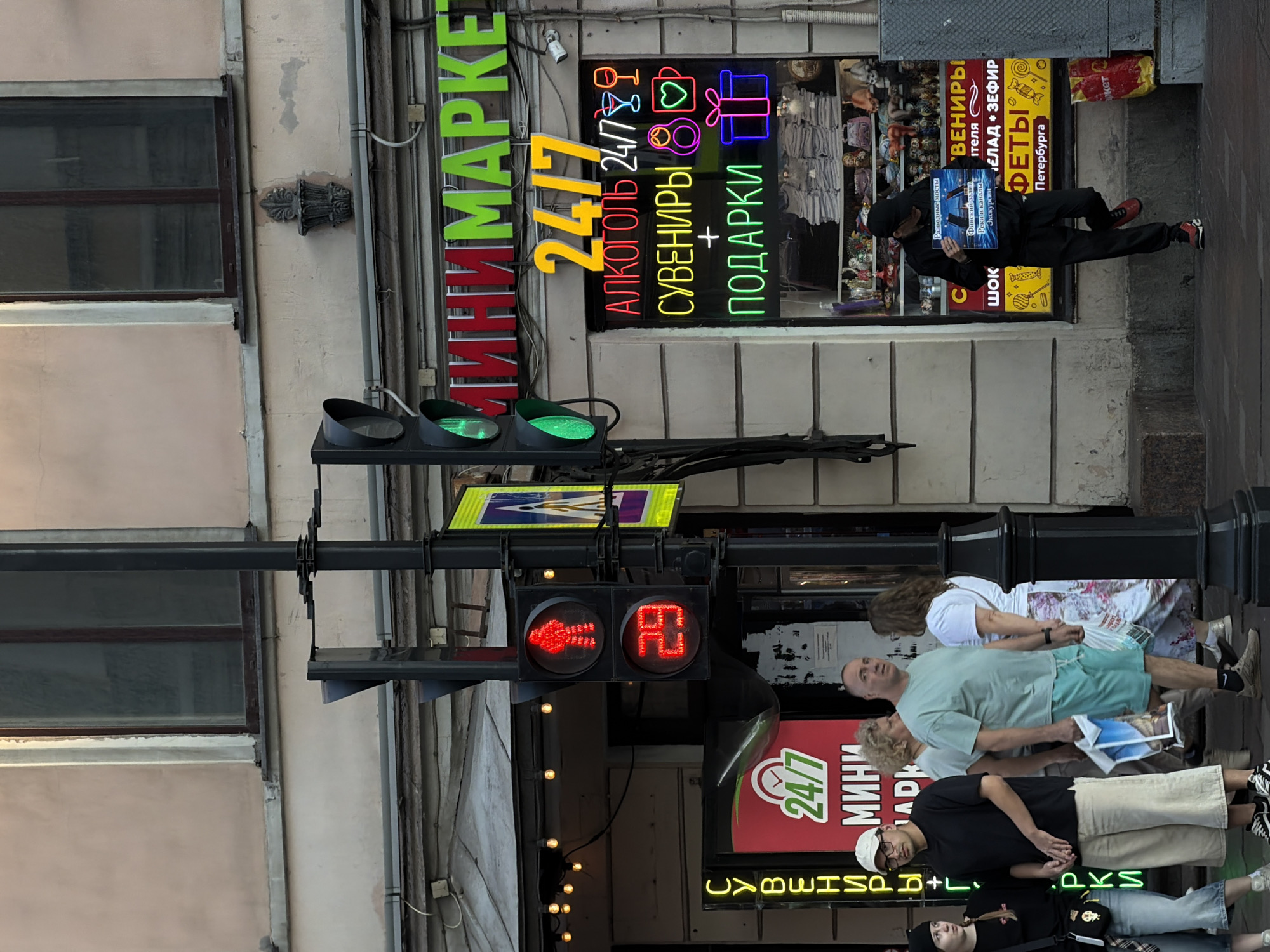 The image depicts a street scene in Saint Petersburg, Russia, near a minimarket. In the foreground, several people are visible. A man wearing a black t-shirt and a white baseball cap stands facing left, holding a folded newspaper. Next to him, a woman with blonde curly hair and a blue t-shirt is also looking to the left. A man in a light blue t-shirt and teal shorts is looking straight ahead, holding a stack of papers. Another woman with long brown hair and a white shirt is partially visible on the right.
A traffic light with a red walking figure and the number "29" illuminated in red is prominent. Above the minimarket entrance, bright neon signs display "МИНИМАРКЕТ 24/7" in red and green. Below this, another neon sign in yellow reads "24/7" and in various colors, lists items like "АЛКОГОЛЬ," "СУВЕНИРЫ," and "ПОДАРКИ." The storefront window displays shelves stocked with various items, including what appear to be souvenirs and gifts.
To the right of the minimarket, a man in black clothing and a black baseball cap is leaning against the building, holding a sign that reads "Разводные мосты, Финский залив, Реки и каналы, Экскурсии." A red and yellow bag is on the ground near him. The building facade is a muted pinkish-beige color, with dark-framed windows. The street is paved with cobblestones or similar material. The overall atmosphere suggests a commercial area during daytime.