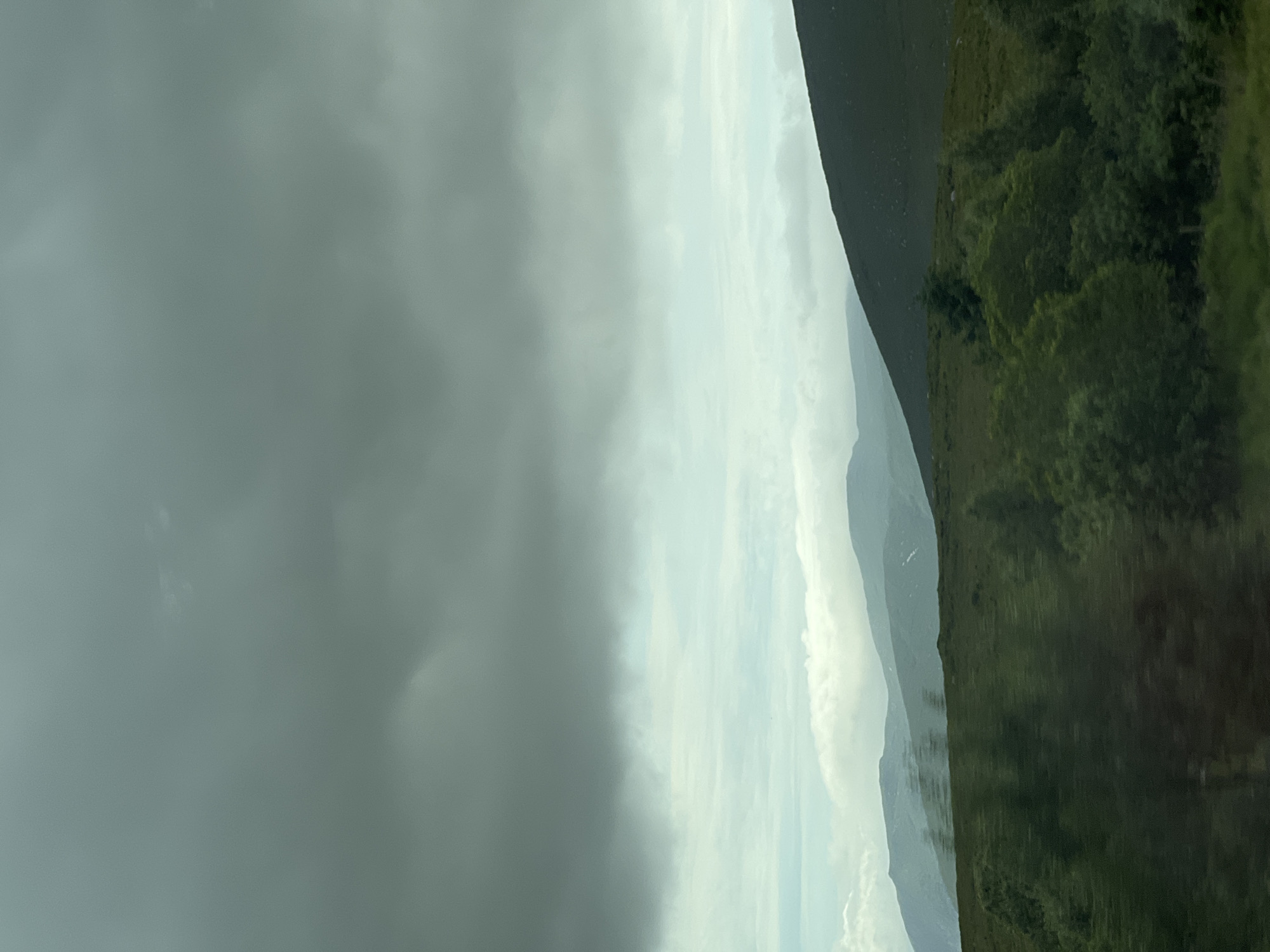 The image depicts a cloudy sky over a vast expanse of rolling green hills. In the foreground, dense foliage and trees blur due to motion, suggesting the photograph was taken from a moving vehicle. The middle ground features a textured green hillside, dotted with smaller shrubs and trees. Beyond this, layers of distant mountains fade into the hazy atmosphere, their slopes covered in muted green and patches of lighter color, possibly indicating sparse vegetation or snow. The sky is dominated by heavy, dark grey clouds, with lighter, diffused clouds and patches of pale blue peeking through in some areas. The overall impression is a moody, atmospheric landscape. The location is identified as Drimnaraw, Ireland. There are no people or specific actions depicted in the scene.