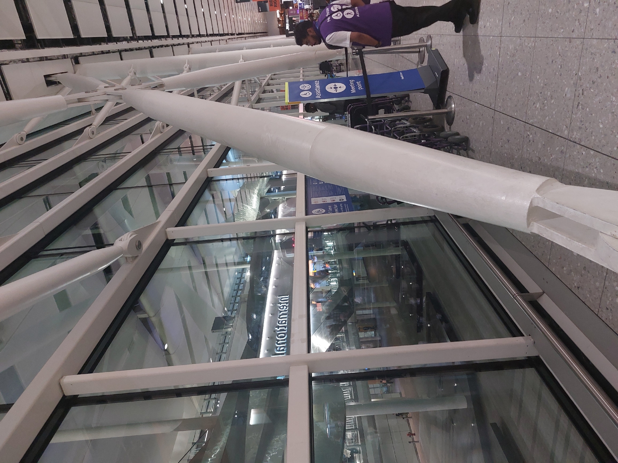 The image depicts an interior scene at an airport, likely Heathrow Airport in London, United Kingdom, given the signage and architectural style. A large glass facade with white structural beams dominates the left side of the frame, reflecting the interior lighting and architecture. Inside the building, a illuminated sign reads "Heathrow." In the foreground, to the right, a person wearing a purple vest with "Heathrow" and an accessibility symbol is visible. They are standing next to a blue sign indicating "Assistance" and "Meeting point," with wheelchairs visible behind them. The flooring is a light-colored tile. The overall atmosphere suggests a busy airport terminal with a focus on passenger assistance.