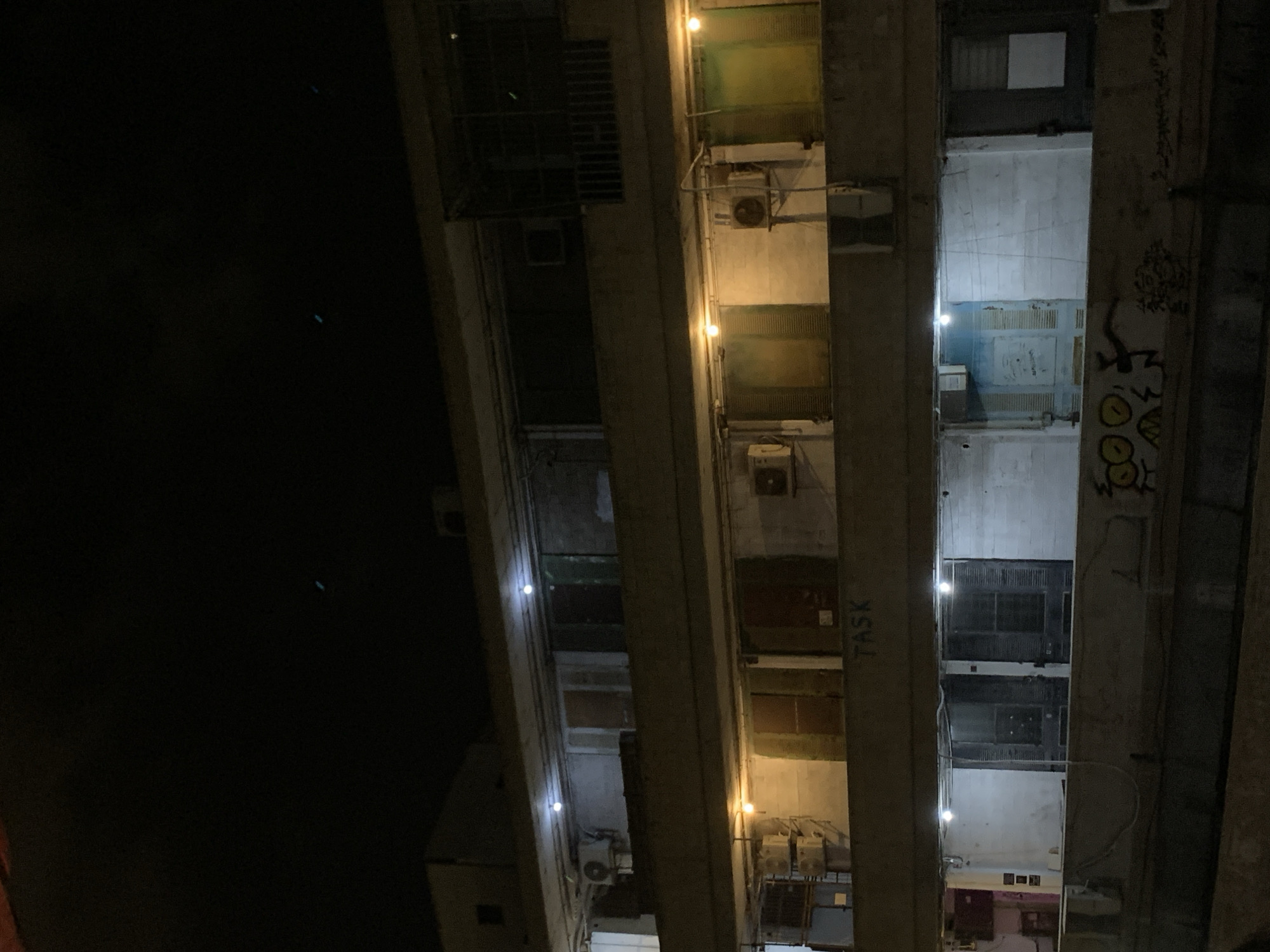An exterior, low-angle night shot captures the facade of a Brutalist-style apartment building in Tel Aviv, Israel. The building features multiple levels with balconies and several windows or doorways, many of which appear to be covered or darkened. Artificial lighting, in the form of exposed bulbs and some interior illumination, casts a warm glow on some sections of the concrete structure. Air conditioning units are visible on some of the balconies, and there are electrical conduits running along the exterior walls. Graffiti, including cartoonish faces and abstract designs, is present on a lower concrete barrier in the foreground. The overall impression is one of urban decay or neglect, with the building appearing aged and somewhat utilitarian.