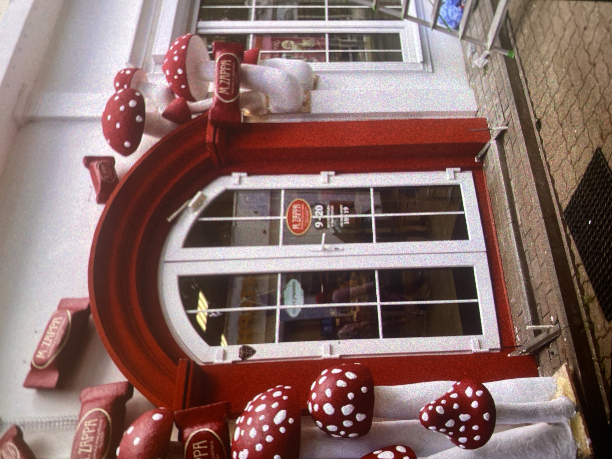 The image captures a whimsical shop facade in Saint Petersburg, Russia. The shop features a distinctive semi-circular red awning over a white-framed glass door with a similarly arched top. The door's glass panes display an interior view, reflecting individuals and objects inside the shop. Decorating the awning and the steps leading to the entrance are red-and-white polka-dotted mushrooms and candy-shaped objects labeled "M. Zappa," adding to the playful, eye-catching design. A window above the shop door also has a white frame. To the right, part of the sidewalk with its tiled surface and a black grate are visible, indicating an urban setting. The photo has a slight tilt, altering the perspective and emphasizing the unusual character of the storefront.