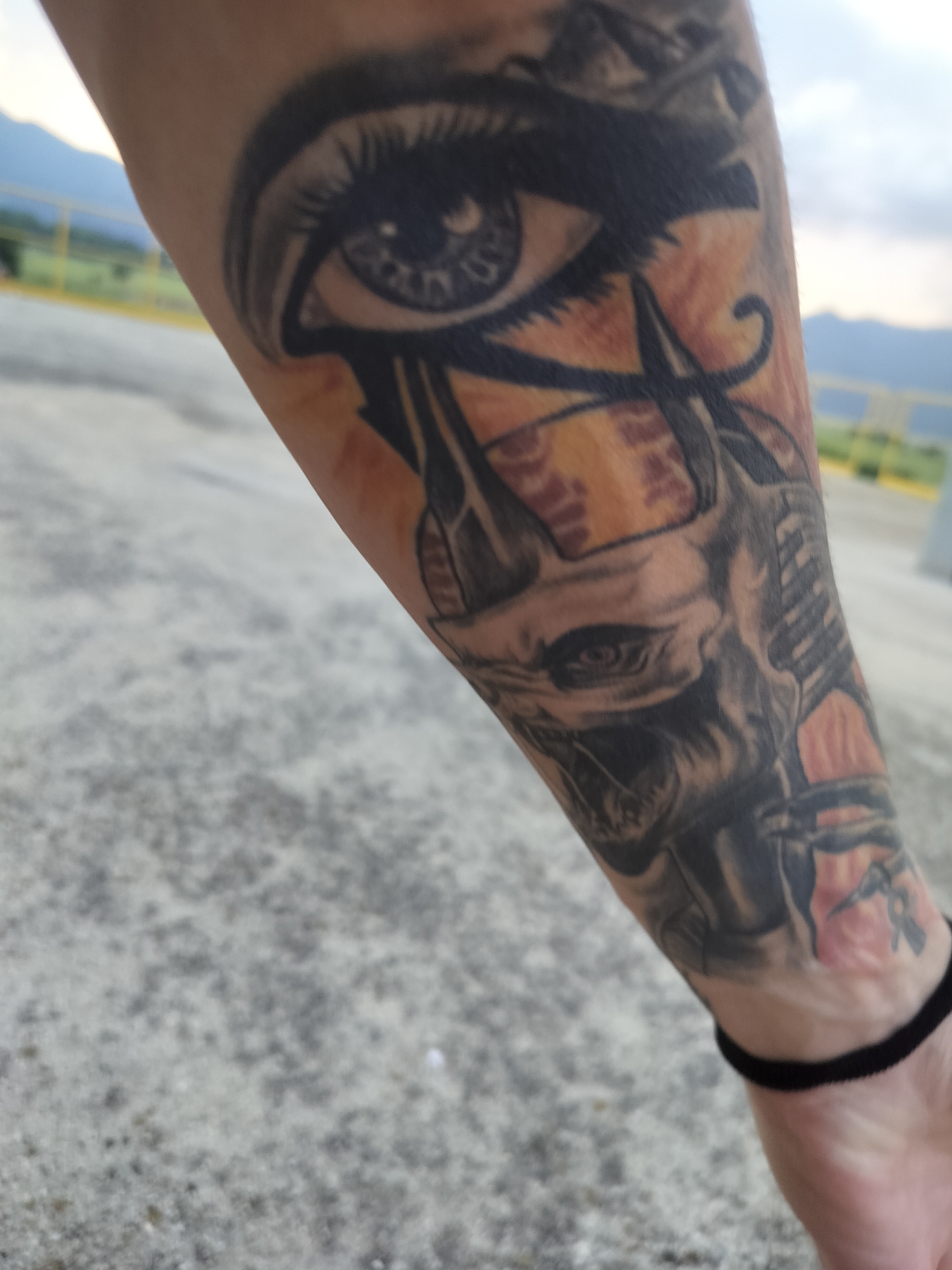The image shows a close-up of a person's tattooed forearm, held outdoors in what appears to be Gorno Sahrane, Bulgaria. The tattoo is a detailed, intricate design predominantly in black and grey with warm orange and brown accents, giving it an ancient Egyptian or mythological theme.
The most prominent elements of the tattoo are a large, stylized eye, reminiscent of the Eye of Horus, positioned towards the upper part of the forearm. Below this, a skull-like or monstrous head with horns is visible, depicted with realistic shading. Further down, near the wrist, parts of the tattoo feature abstract patterns and what appear to be hieroglyph-like symbols or decorative elements. The background of the tattoo incorporates textured areas with red and orange hues. The person is wearing a simple black wristband on their wrist.
The arm is held against a blurred outdoor background. The ground is a grey, textured paved surface, possibly a road or parking lot. In the mid-ground, there are blurred green landscapes and a yellow or light-colored railing. Distant, hazy mountains are visible under a bright, overcast or partly cloudy sky, suggesting the time of day is either late afternoon or early evening. The overall lighting is soft and diffused. No specific activity is captured other than the display of the tattoo. There is no discernible text within the image.