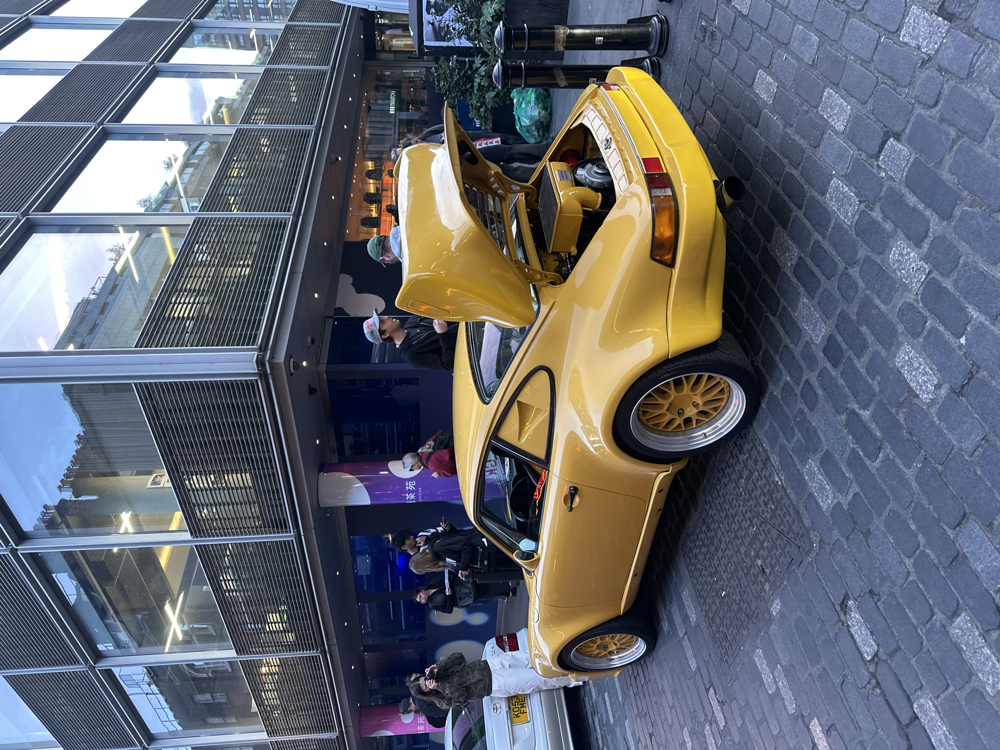 A vibrant yellow classic Porsche 911 is prominently displayed on a cobblestone street in London, United Kingdom. The car's rear engine cover is open, revealing components of its air-cooled engine, and it is fitted with distinctive gold-colored, multi-spoke wheels with polished rims.
Several individuals are gathered around the vehicle. One man wearing a green cap and another in a white cap are observing the exposed engine. A woman in a camouflage jacket and white pants is actively photographing the Porsche with a camera. Other people are visible in the background, some looking at the car, others simply walking past.
The setting features modern urban architecture, with large glass-paned buildings reflecting the surroundings. These buildings have prominent horizontal slat designs. Black bollards are visible along the street, and a green plastic bag rests nearby. Behind the Porsche, a white car, possibly a Toyota, is partially visible, displaying a UK license plate that reads "KO53 PME." Further in the background, a sign partially reads "FEEL MATCHCHA" with additional text in an Asian script on a purple and white pillar.
The scene suggests an informal car display or a car spotting event, with enthusiasts admiring the classic sports car. The time of day appears to be daytime, possibly overcast, given the lighting conditions. The overall atmosphere is one of casual interest and appreciation for the unique automobile.
