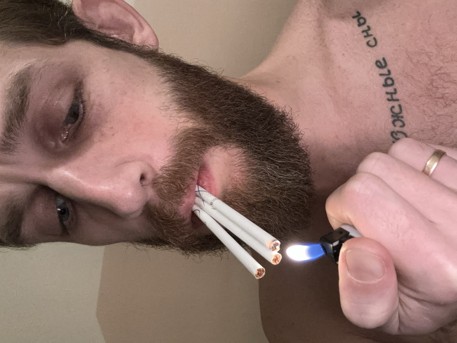 The image presents a close-up, angled view of a man, likely indoors in Saint Petersburg, Russia. His face is visible from the forehead down, with his head tilted, showing his left eye looking downwards. He has a full brown beard and what appear to be light-colored eyes.
The central activity is the man lighting multiple cigarettes at once. Three cigarettes are held between his lips, and his right hand holds a lighter with a distinct blue flame, positioned to ignite the tips of the cigarettes. A plain gold wedding band is visible on one of the fingers of his right hand.
A notable detail is a tattoo on his upper chest or shoulder area, written in Cyrillic script. The text reads "Божные сны," which translates to "Divine dreams" or "Godly dreams." The background is a plain, light-colored surface, suggesting an indoor setting. The lighting is somewhat dim, contributing to a slightly somber or focused mood. The man's expression is concentrated on the act of lighting the cigarettes.