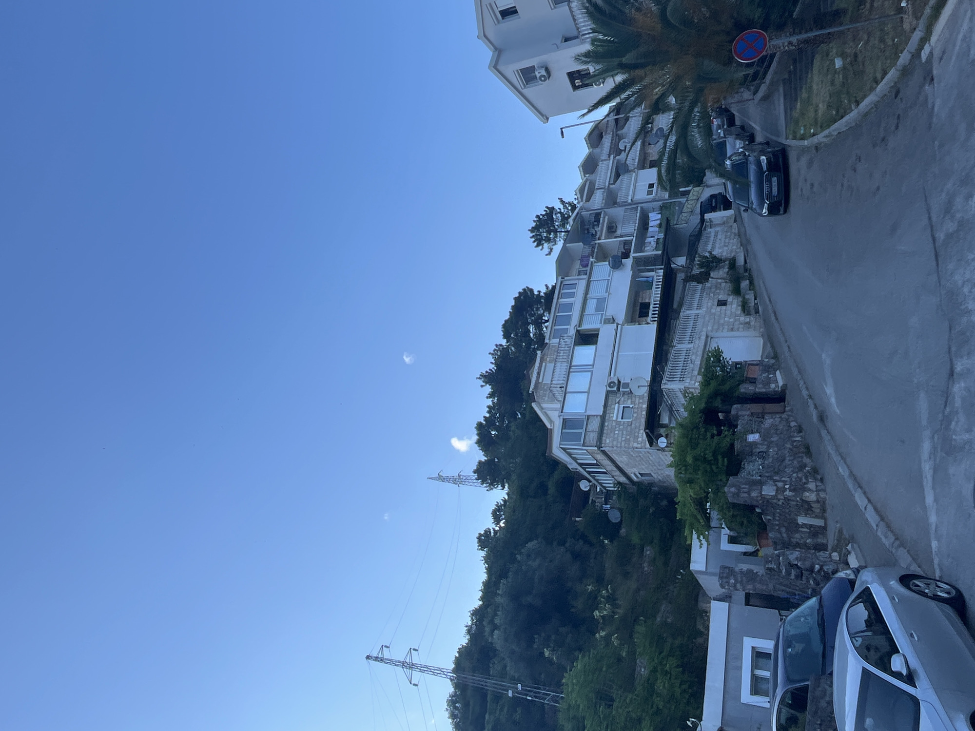 The image captures a residential street in Budva, Montenegro, characterized by its hilly terrain. On the left side of the street, a multi-story building with white facades and numerous balconies is visible, nestled against a lush green hillside. Power lines stretch across the sky, supported by tall metal towers, indicating the presence of significant electrical infrastructure.
The street itself is paved and curves upwards, with several cars parked along its side. The vehicles appear to be modern sedans, one of which is a dark-colored Audi. The buildings lining the street are predominantly white, with some exhibiting stone detailing. Balconies are a common feature, some adorned with laundry lines.
To the right of the street, a tall palm tree stands, its fronds reaching towards the clear blue sky. A red and blue circular sign with a horizontal bar is affixed to a pole near the palm tree, likely a traffic or parking regulation sign. The sky is vast and clear, with a hint of a wispy cloud in the distance. The overall impression is of a coastal town in a Mediterranean climate, with architecture adapted to the sloped landscape and a blend of natural greenery and urban development.