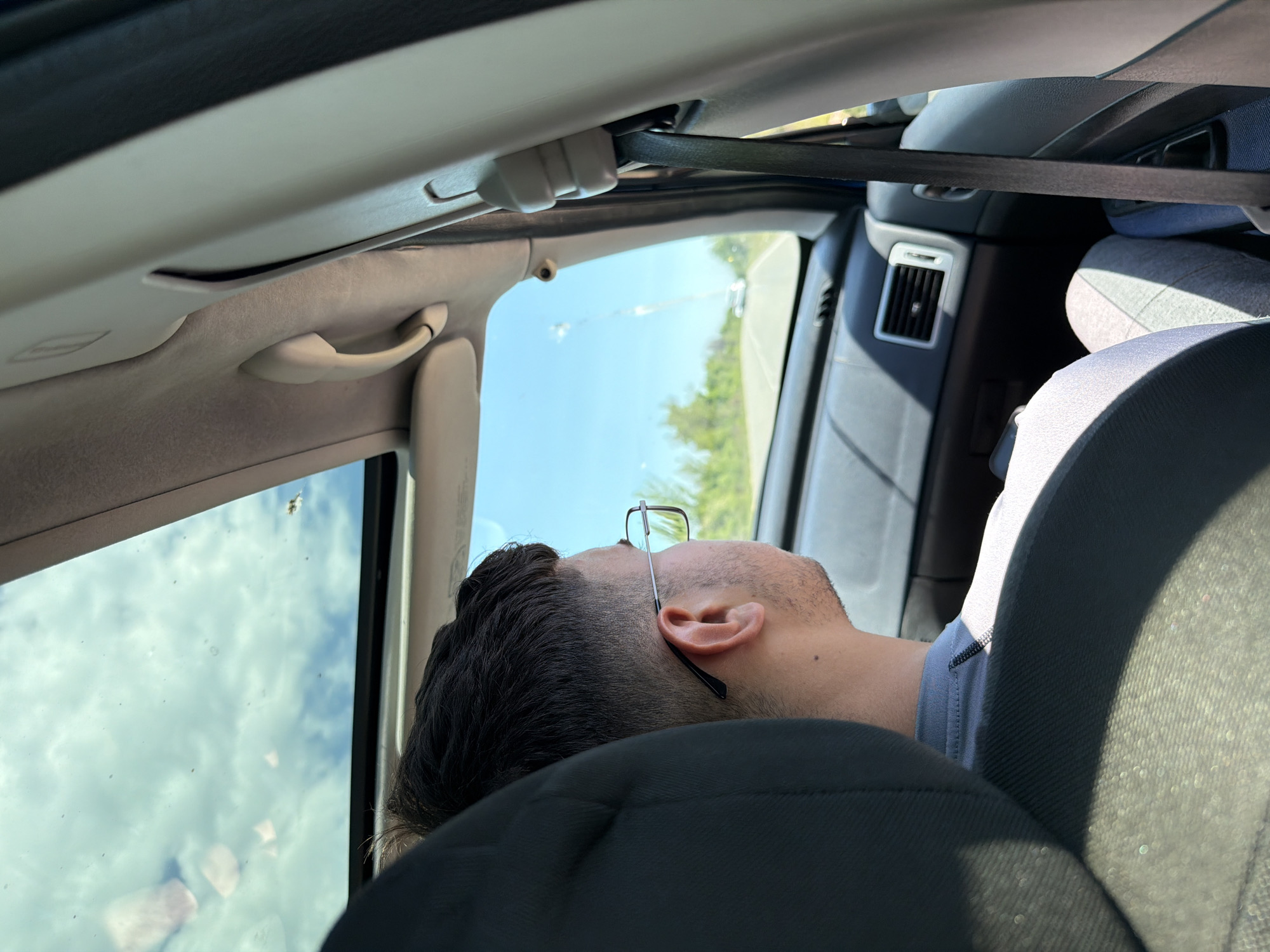 The image shows a person lying down in a car. The person is wearing glasses and has dark hair. The person's head is resting on a black car seat. A seatbelt is visible. The car's interior is visible, including the roof, handle, and air vent. Outside of the car is a blue sky, trees, and the edge of a road. There is no information to determine if the person is in Belovitsa, Bulgaria.