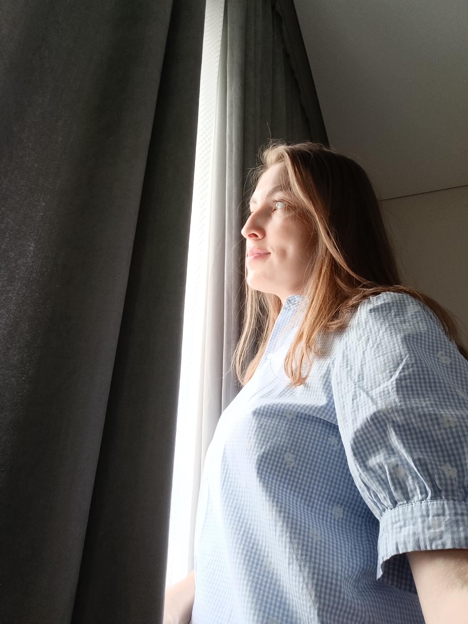 A fair-skinned woman with light brown hair is looking out of a window. She has a slight smile and is wearing a blue and white checkered blouse with puffy sleeves. She is positioned in front of a window with light sheer curtains and heavy dark grey drapes. The location is an interior space, likely a room, with a view of the outside, as suggested by the light streaming in. No additional context is provided to identify its location in the city of Chaikovsky, Russia.