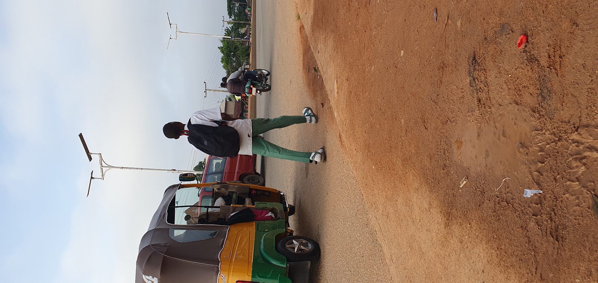 In Bukuru, Nigeria, a young male wearing a white short-sleeved shirt, green trousers, and sandals walks across a paved road. He carries a black backpack on his shoulders. To his left, a yellow and green auto-rickshaw is parked. In the background, a red van and a motorcycle carrying two people are visible. The scene is set against a cloudy sky with streetlights present on the roadside. The foreground consists of a dirt road with some puddles and debris, including a red object that appears to be a bottle cap.