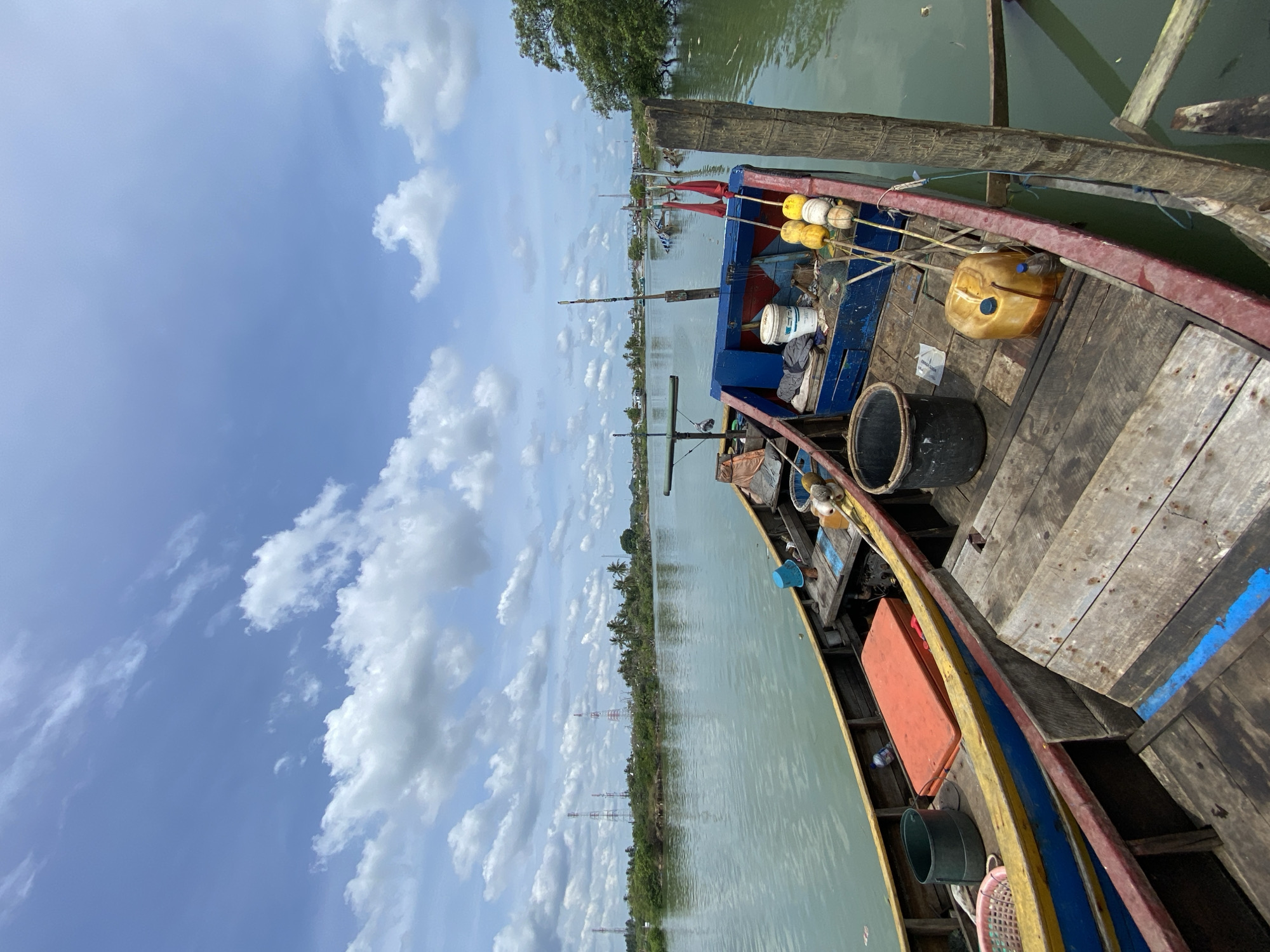 The image shows the interior and immediate surroundings of a moored wooden fishing boat. The boat is located on a river or canal near Tanjongkapai, Indonesia, as indicated by the presence of communication towers and coastal vegetation in the background. The boat's hull is painted with red, yellow, and blue stripes. On the deck are various items, including a yellow fuel container, a black bucket, white buoys, and what appear to be fishing supplies. The water is a murky green color. The sky is blue with puffy white clouds. The boat is tied to a wooden post or pier.