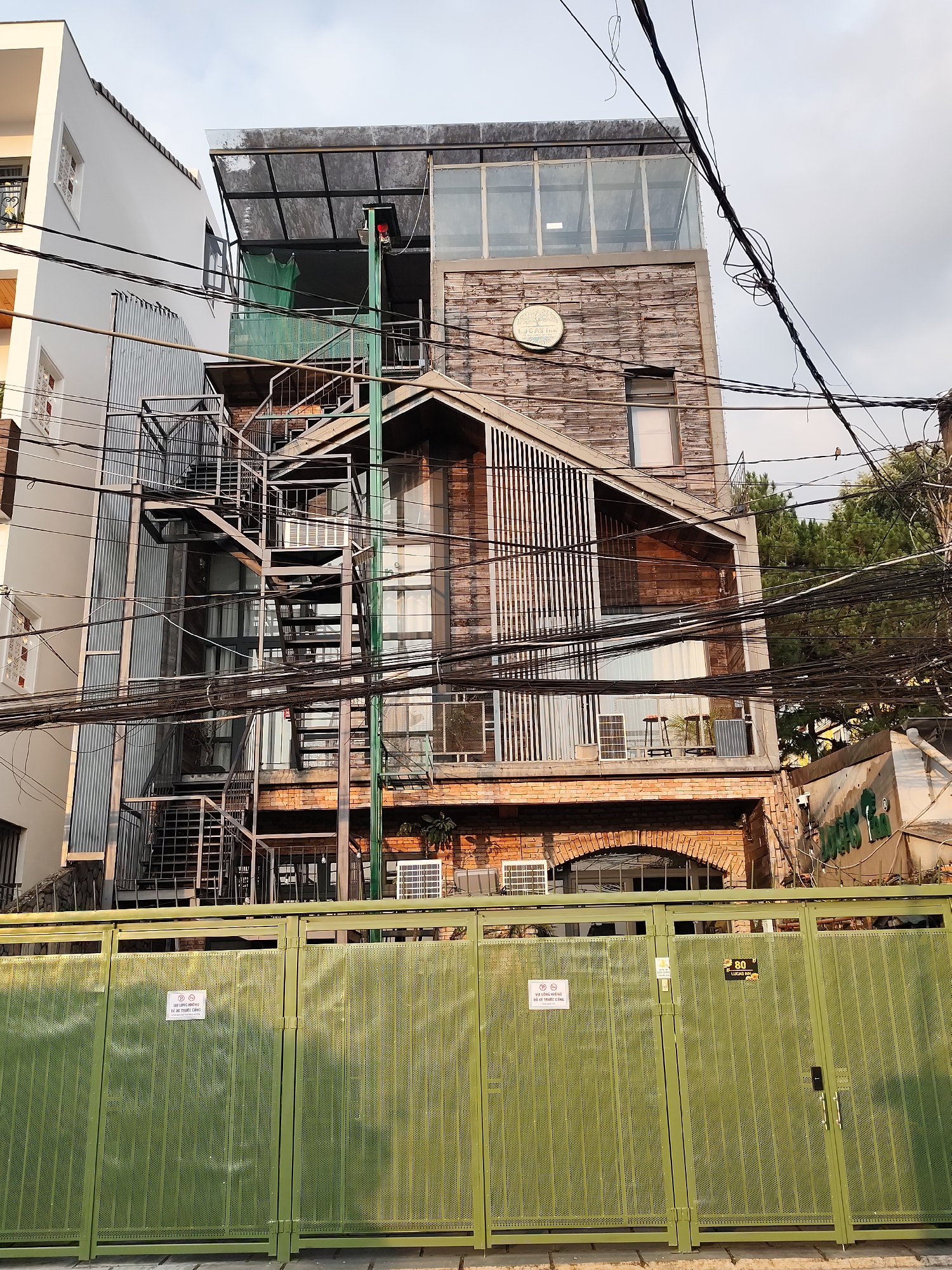 The image depicts a multi-story building in Da Lat, Vietnam, situated behind a green metal gate. The building is characterized by a diverse architectural style, with brickwork at the base, a wooden facade, a roof with vertical wooden slats, and a glass-enclosed upper level. A metal spiral staircase runs along the exterior of the building. Numerous electrical wires crisscross the foreground and obscure parts of the building. Signage is visible on the gate, including the text "VUI LÒNG KHÔNG ĐỂ XE TRƯỚC CỔNG" (please do not park in front of the gate). Also, a sign with "LUCAS Inn" and the number "80" can be seen. The buildings are set against a cloudy sky.