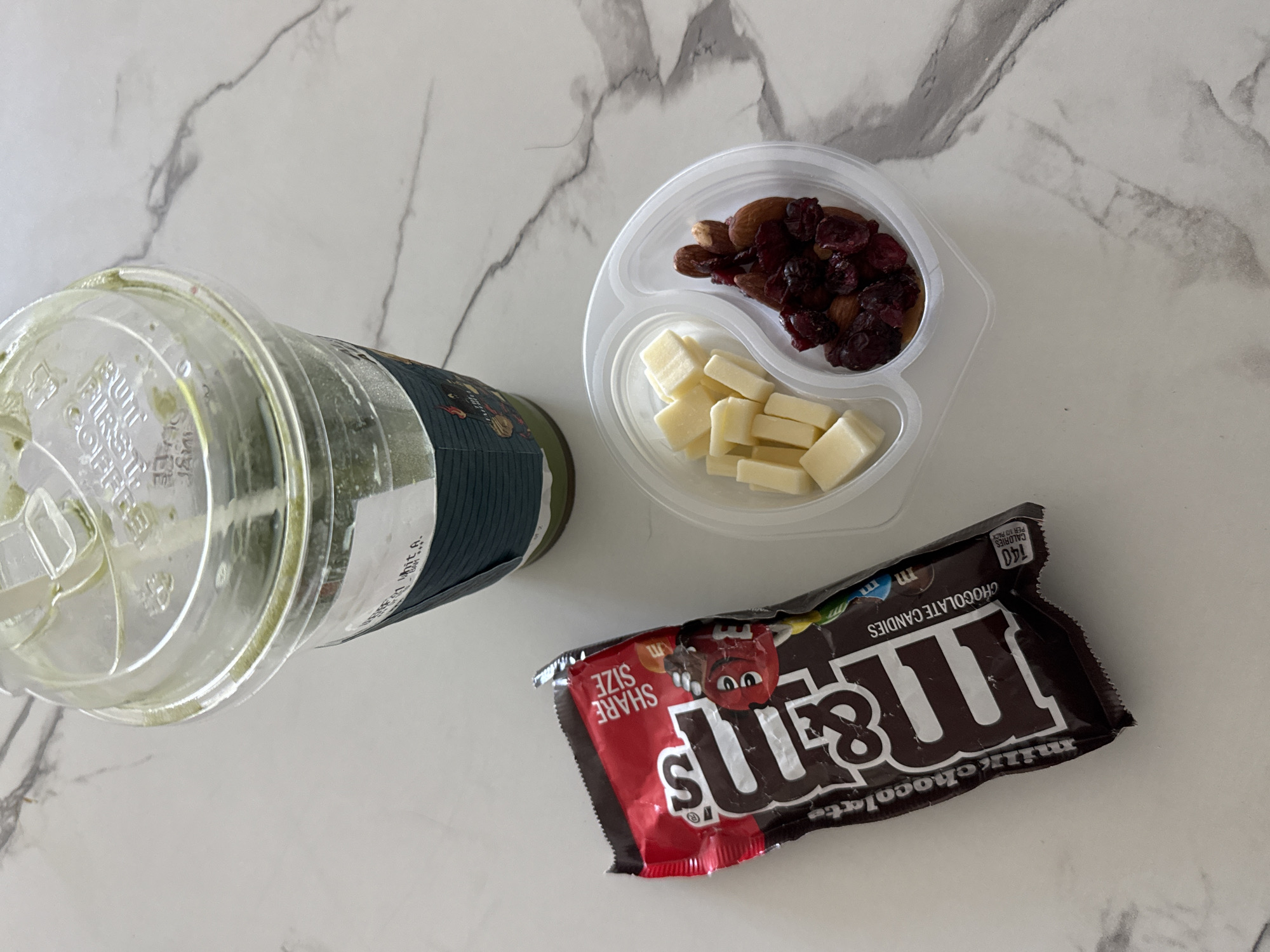 The image shows a flat lay of various snacks and a drink on a light-colored, possibly marble, countertop.
From left to right, there is a plastic cup with a lid, likely containing an iced matcha latte. Next to the drink is a clear plastic container with two compartments: one filled with cubes of white cheese and the other with dried cranberries and almonds. To the right of the plastic container is a share-size bag of Milk Chocolate M&M's, partially opened.
There are no people visible in the image. The environment appears to be indoors, on a kitchen counter. The location is not provided in the image.
