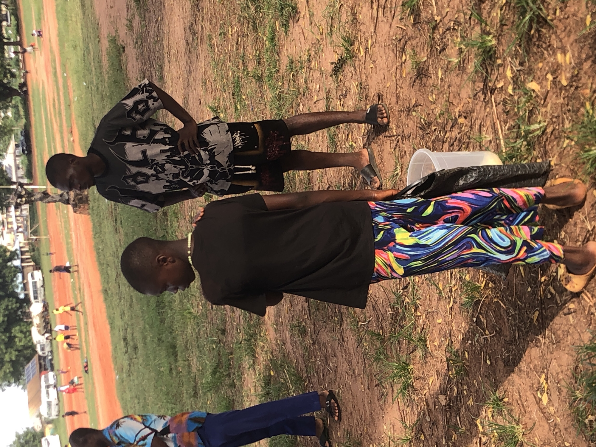 The image depicts an outdoor scene in Ukwu-Abwa, Nigeria, featuring several individuals on what appears to be a dry, reddish-brown dirt and grassy field.
In the immediate foreground, two young men are the primary subjects. The man on the right is dressed in a short-sleeved shirt with a grey, black, and white abstract pattern, black shorts featuring a subtle design, and black slide sandals. He stands with one hand on his hip, looking towards the left side of the frame. Closer to the center, a second man is seen from behind, wearing a plain black t-shirt and eye-catching pants with a vibrant, swirling abstract pattern in shades of blue, pink, yellow, and green. He wears light brown sandals. On the ground near his feet, a translucent white plastic bucket and a black bag are visible. A third individual is partially captured in the bottom-left corner, wearing a blue patterned shirt, blue pants, and black sandals.
In the mid-ground and background, a wider expanse of the field is visible, including a prominent reddish-brown track or path running alongside grassy areas. Several other people are scattered across this area, some appearing to be walking or standing, suggesting a casual gathering or a local sports event might be taking place. The scene is brightly illuminated, indicating a clear, sunny daytime setting. There is no visible text in the image. The overall atmosphere appears relaxed and informal.