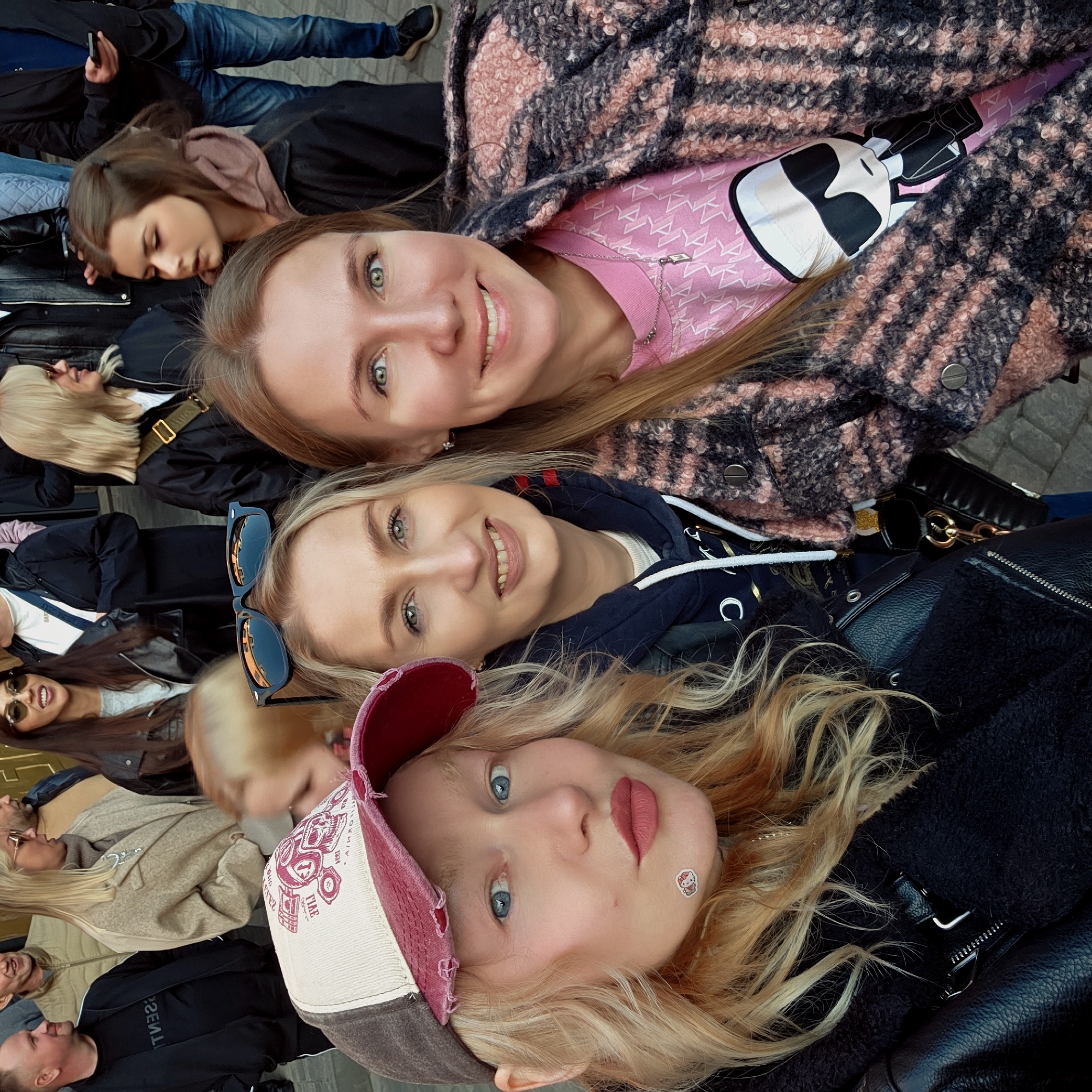 A close-up shot portrays a group of women lying on the ground in an urban setting, possibly in an unidentified city. Three women are prominently featured: one with red-blonde hair wearing a baseball cap, another with blonde hair and sunglasses placed atop her head, and a third with light brown hair. The woman with light brown hair is wearing a pink t-shirt with a white and black design under a pink and gray patterned coat. Behind them, a diverse group of people are either seated on benches or lying down, some of whom are wearing jackets. One individual holds a phone. The ground beneath them appears to be paved.