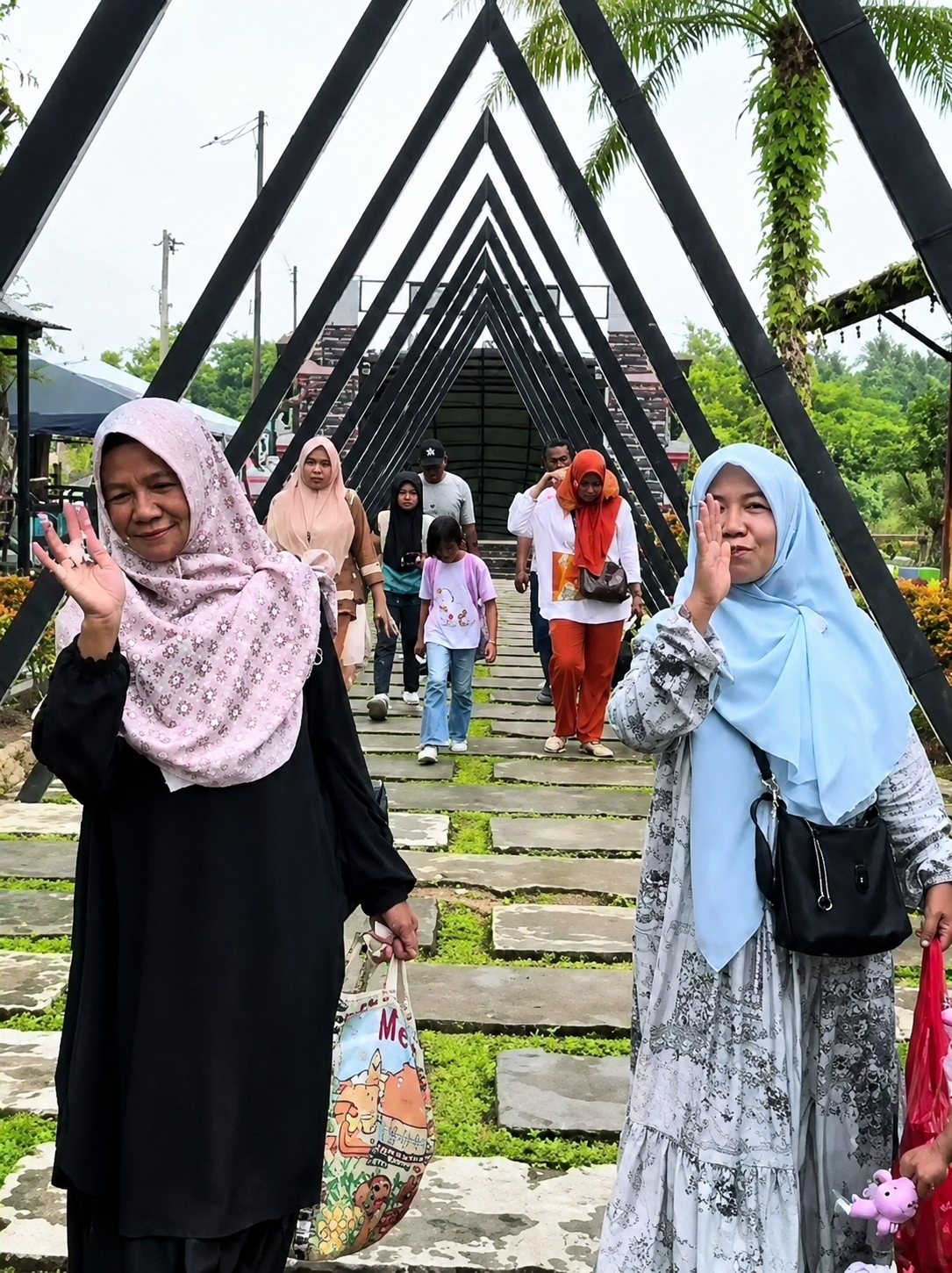 In Eampat Kwalamencirim, Indonesia, two women in hijabs are prominently featured in the foreground of a pathway lined with stone slabs and lush green vegetation. The woman on the left, wearing a black abaya and a light pink hijab with a floral pattern, waves to the camera with her right hand. She carries a colorful tote bag with cartoon characters. The woman on the right wears a light blue hijab and a grey and white patterned dress, also carrying a black shoulder bag. She is also in motion, her right hand raised. Behind them, a group of people, including children and adults, are walking away from the camera down a covered walkway constructed with angled black metal beams, forming a pointed archway. The background reveals more greenery and hints of structures, suggesting an outdoor recreational area or park. The sky is overcast, and the lighting is diffused.
